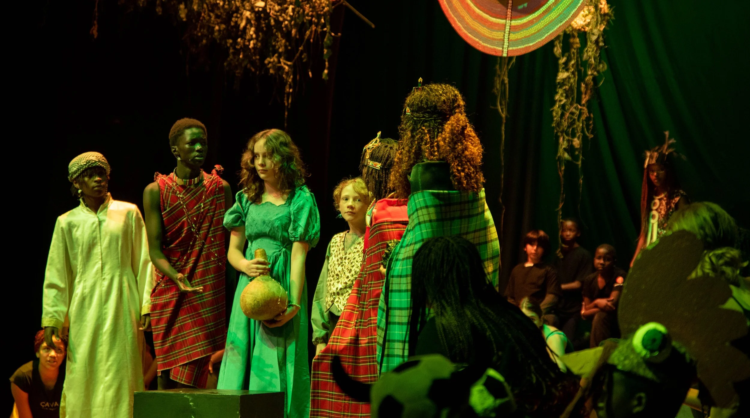 Children on stage during a theatrical performance with costumes and props, some dressed in traditional African attire, with a dark background and vibrant lighting.
