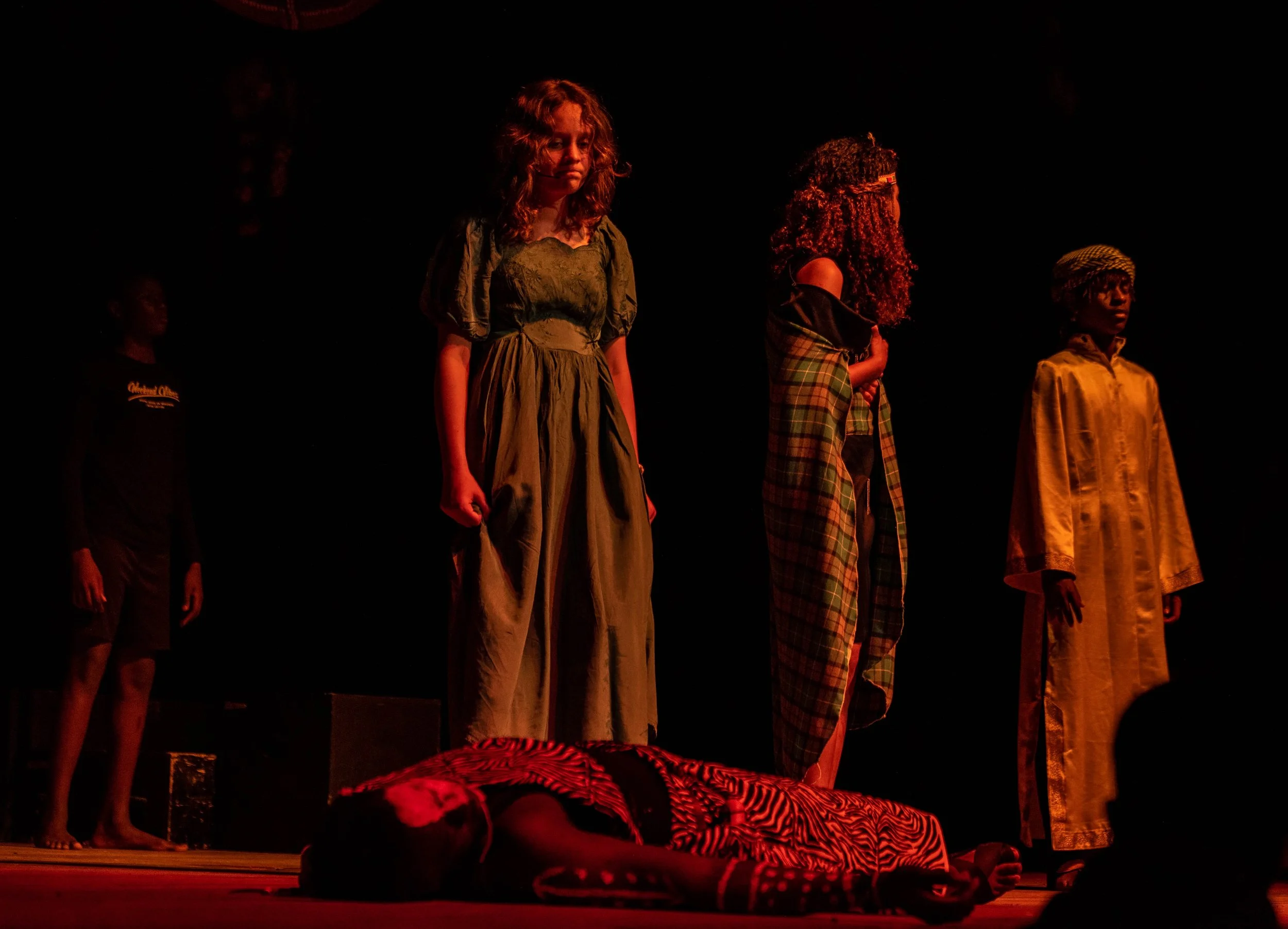 Four children standing on stage in theatrical costumes, one is lying on the floor in a zebra-patterned costume. The background is dark, with stage lighting highlighting the children.