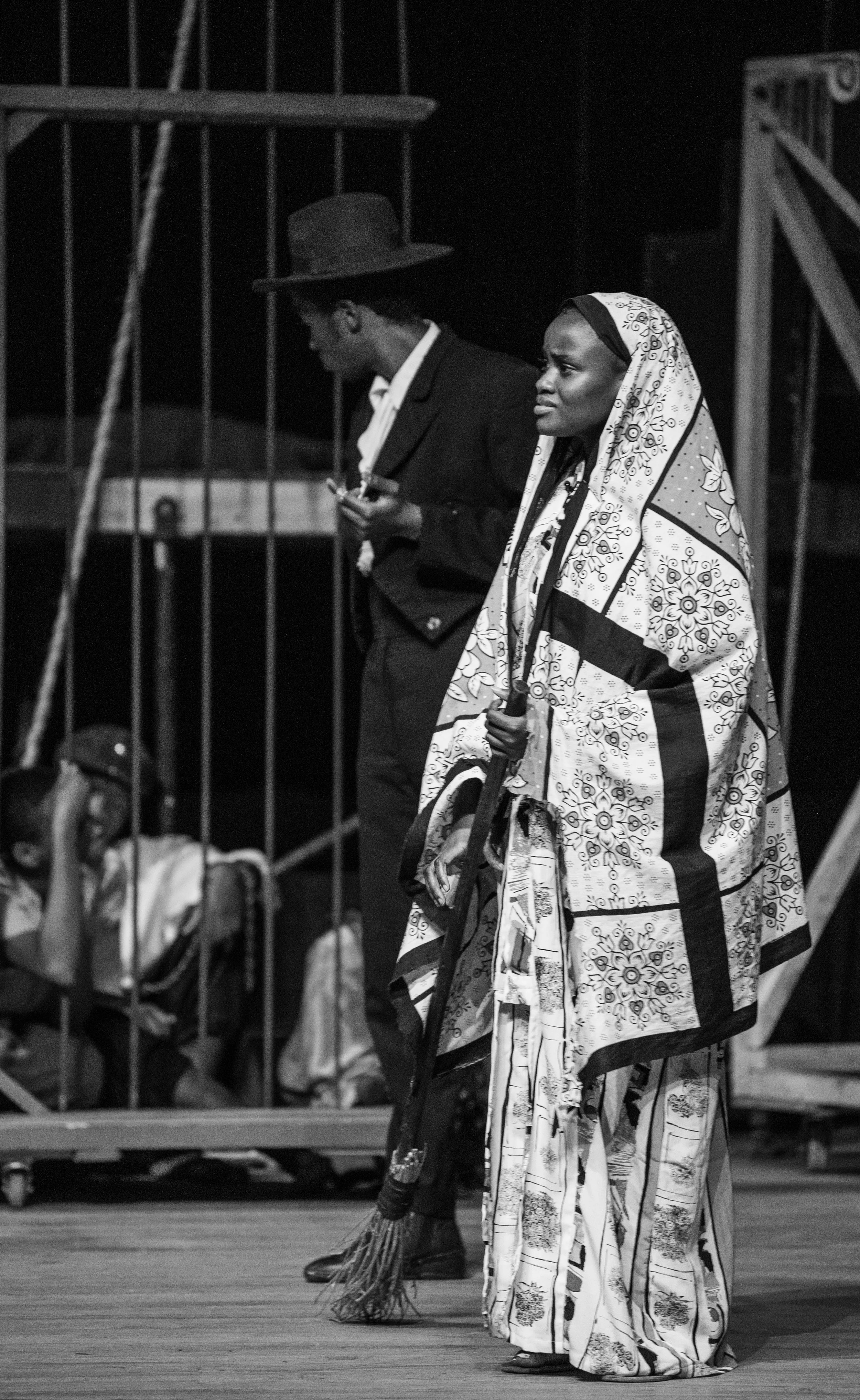 A woman in traditional attire, holding a rifle, standing on stage with a somber expression. A man in justice attire stands behind her, looking down. A person in casual clothes is sitting on the floor in the background, behind a fence.