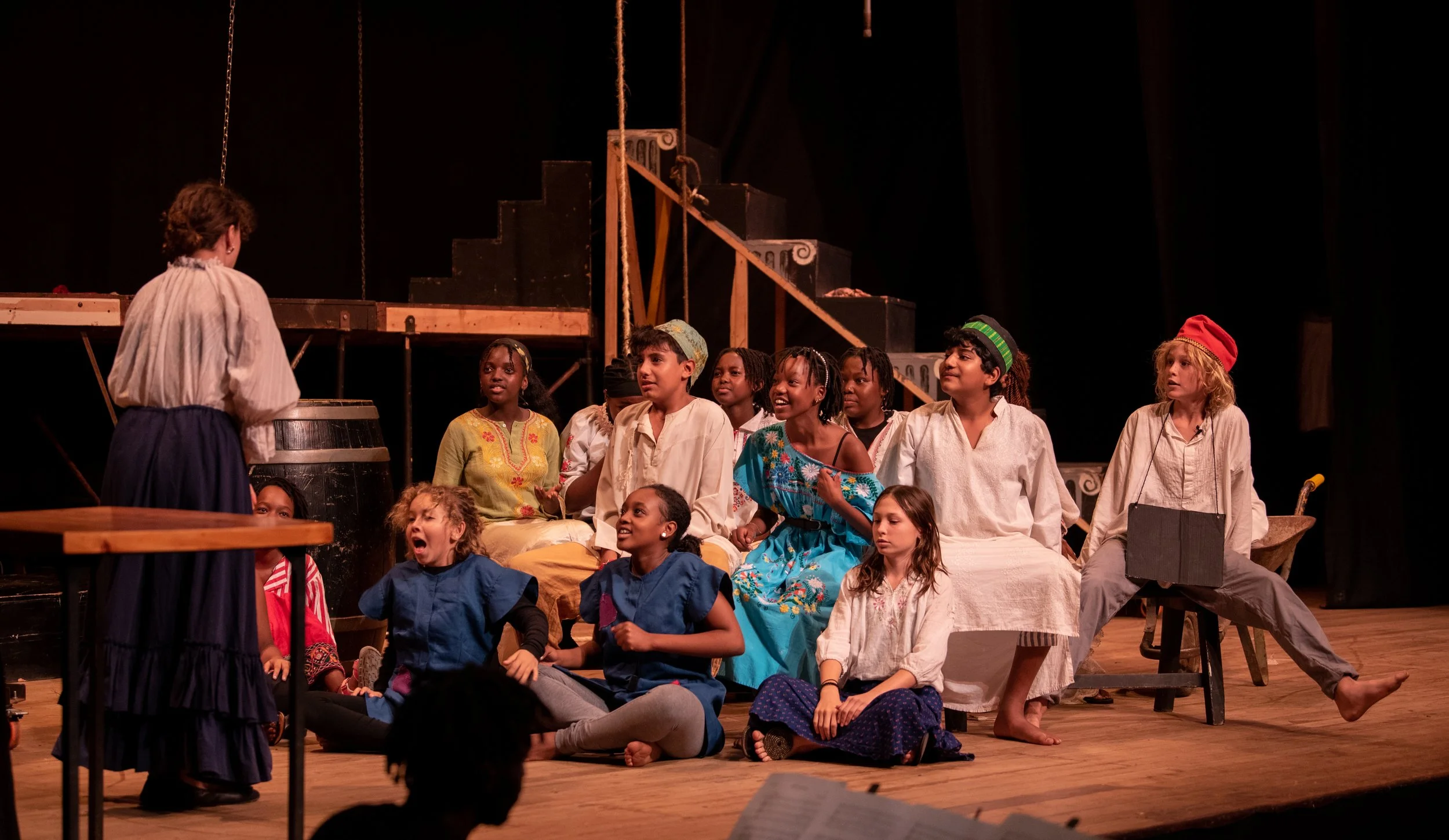 Children on stage performing in a play, sitting and standing in front of a dark backdrop with set pieces, dressed in costumes resembling a historical or fairy tale setting.