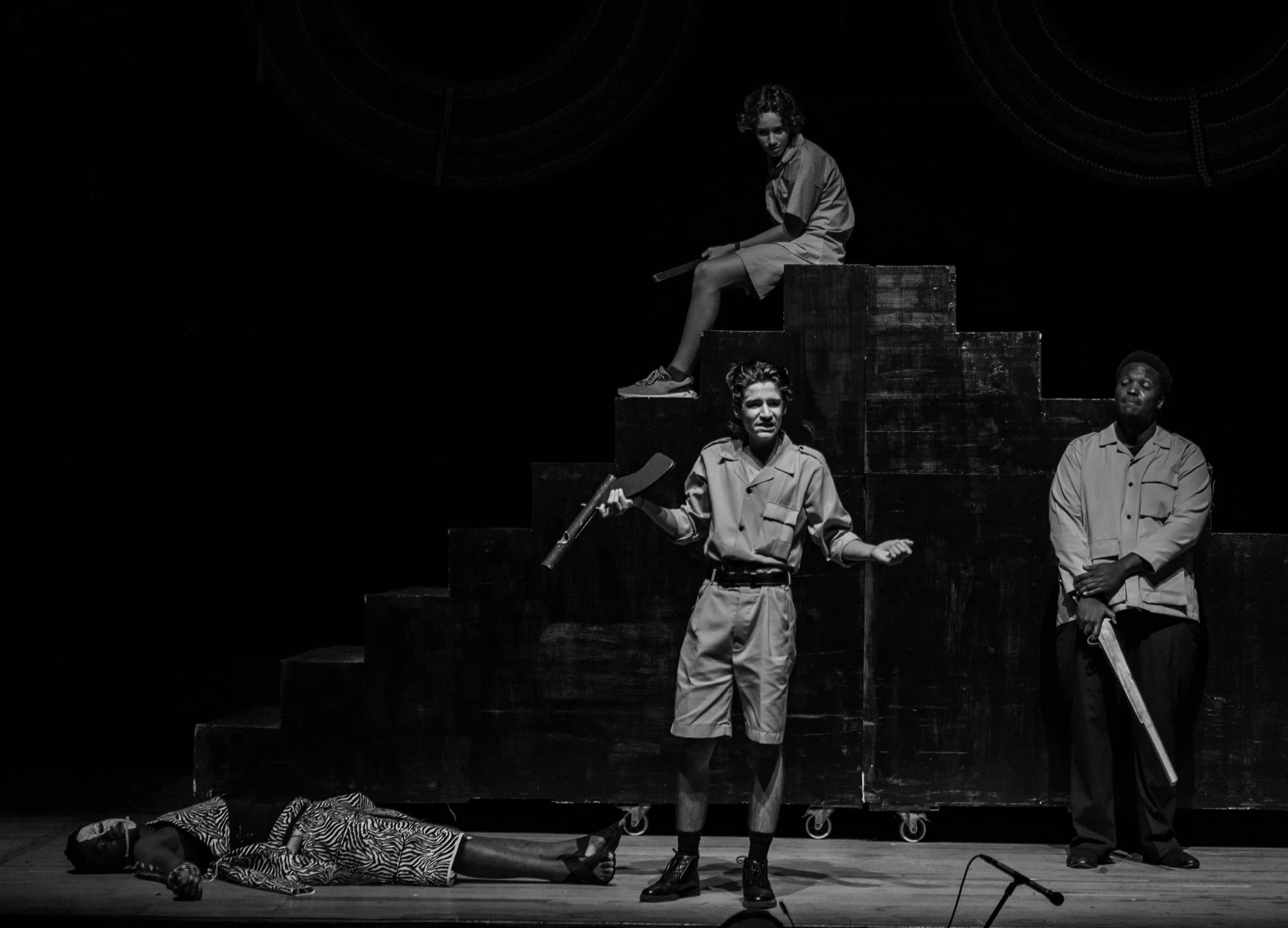 Black and white photo of four actors on stage performing a play, with three standing and one lying on the floor, against a dark background with large circular designs.