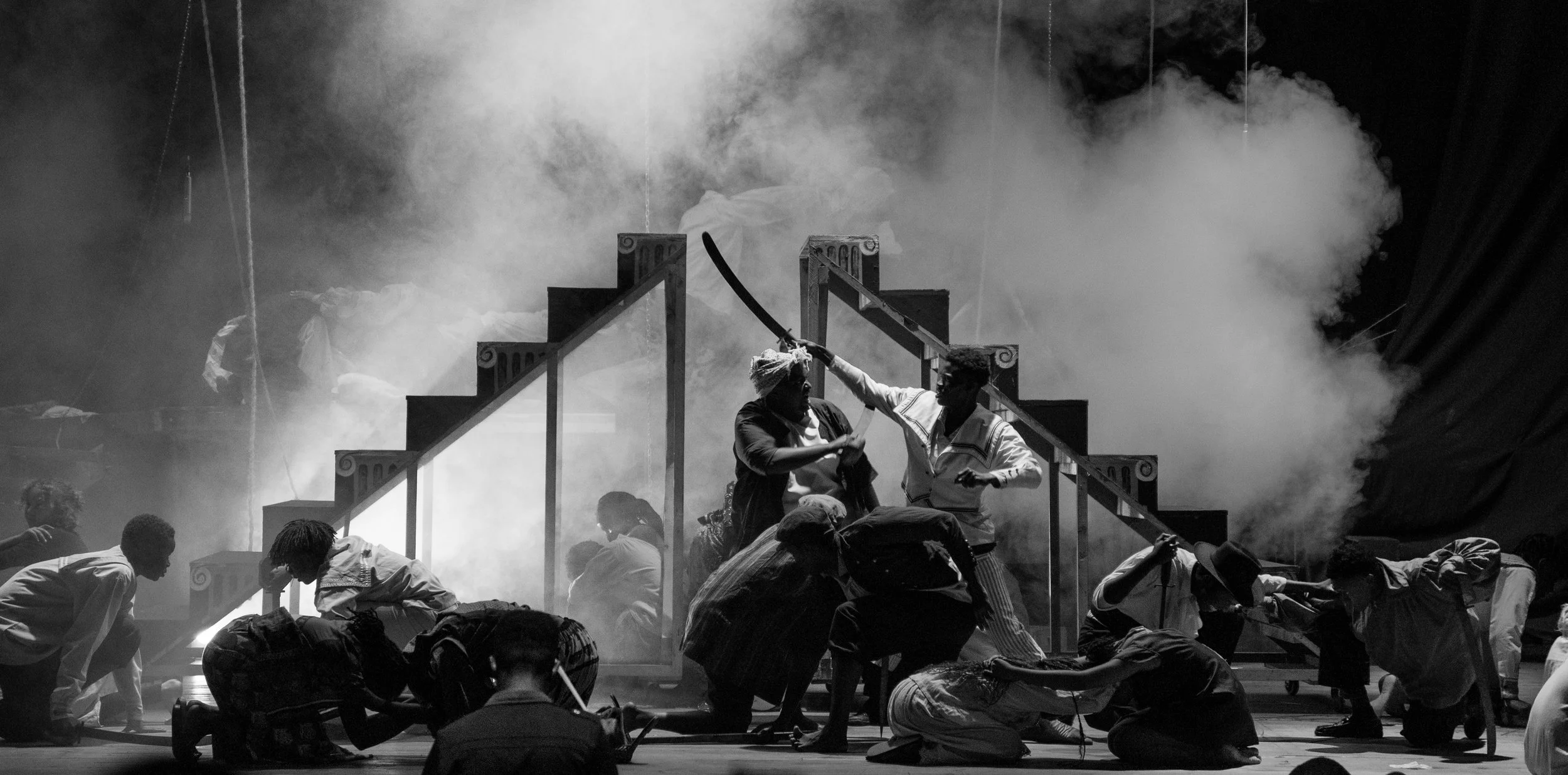 The scene appears to be a theatrical or stage performance with smoke and a group of actors. Two actors are engaged in a dramatic confrontation, with one holding a curved prop, possibly a sword or whip. Other actors are kneeling or crouching on stage,