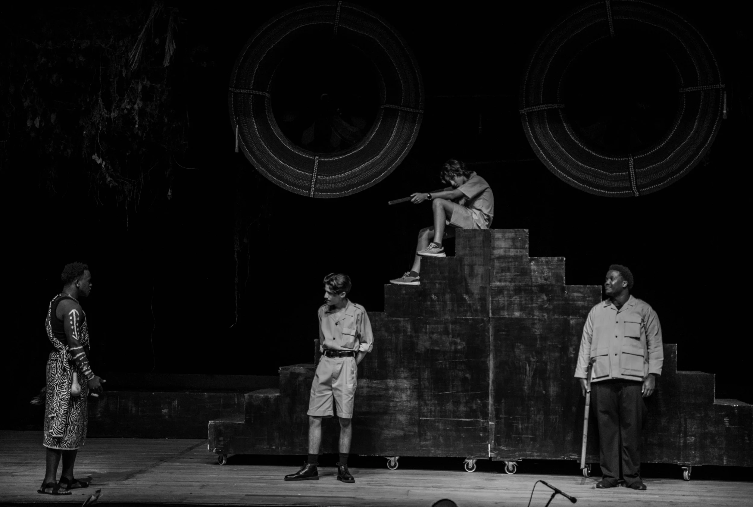 Four children perform on a stage with a set resembling steps. One child sits on top, aiming a toy gun. Two children stand on the stage, one facing the girl, and the other with a cane. The background features large circular props.