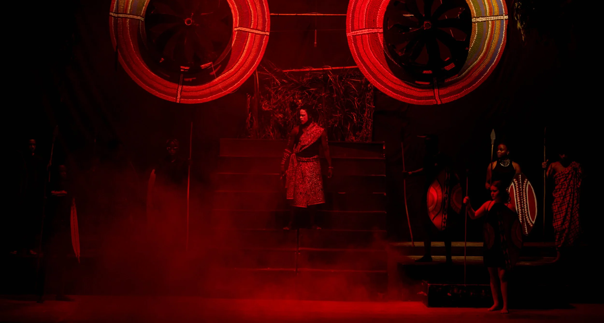 The image depicts a theatrical stage performance with several performers dressed in traditional African attire. One woman stands at the center on a raised platform, while others are positioned around her holding shields and spears. The stage is illum