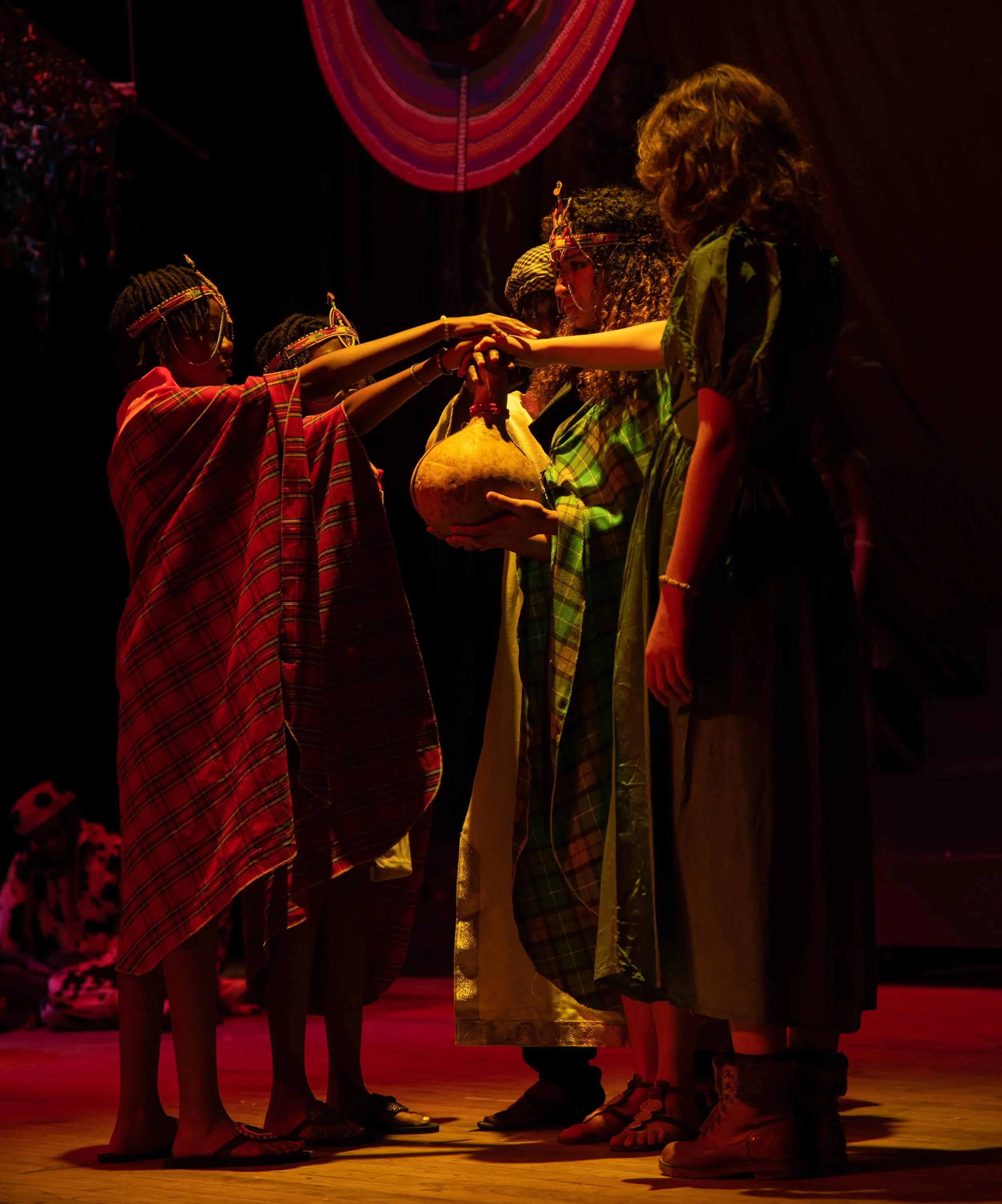 Group of children on stage during a performance, some dressed in traditional clothing, with a dark background and stage lighting.