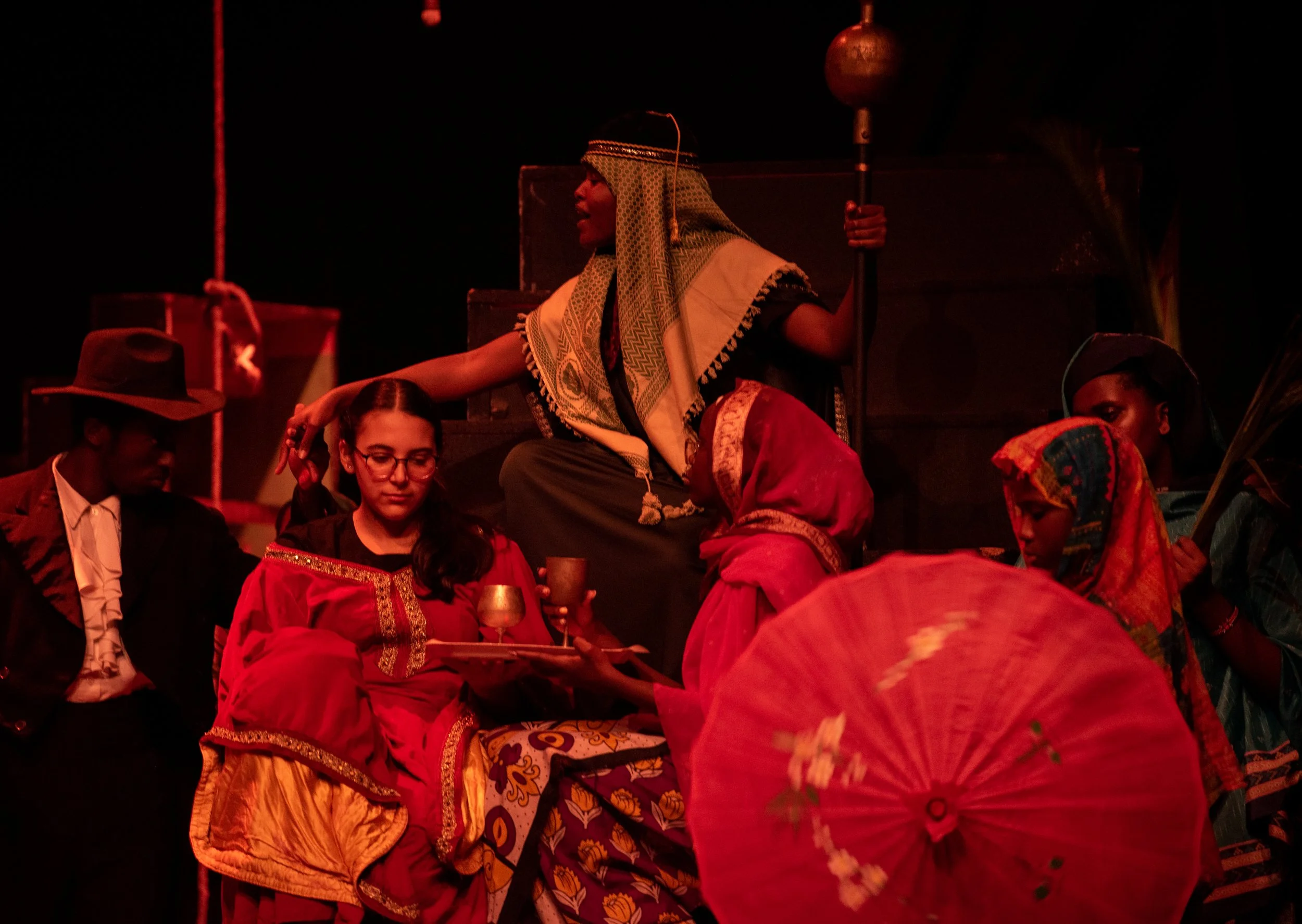 Actors in colorful costumes performing a theatrical scene, with some seated and others standing, including a woman holding a staff and a sun-shaped ornament, set against a dark background.