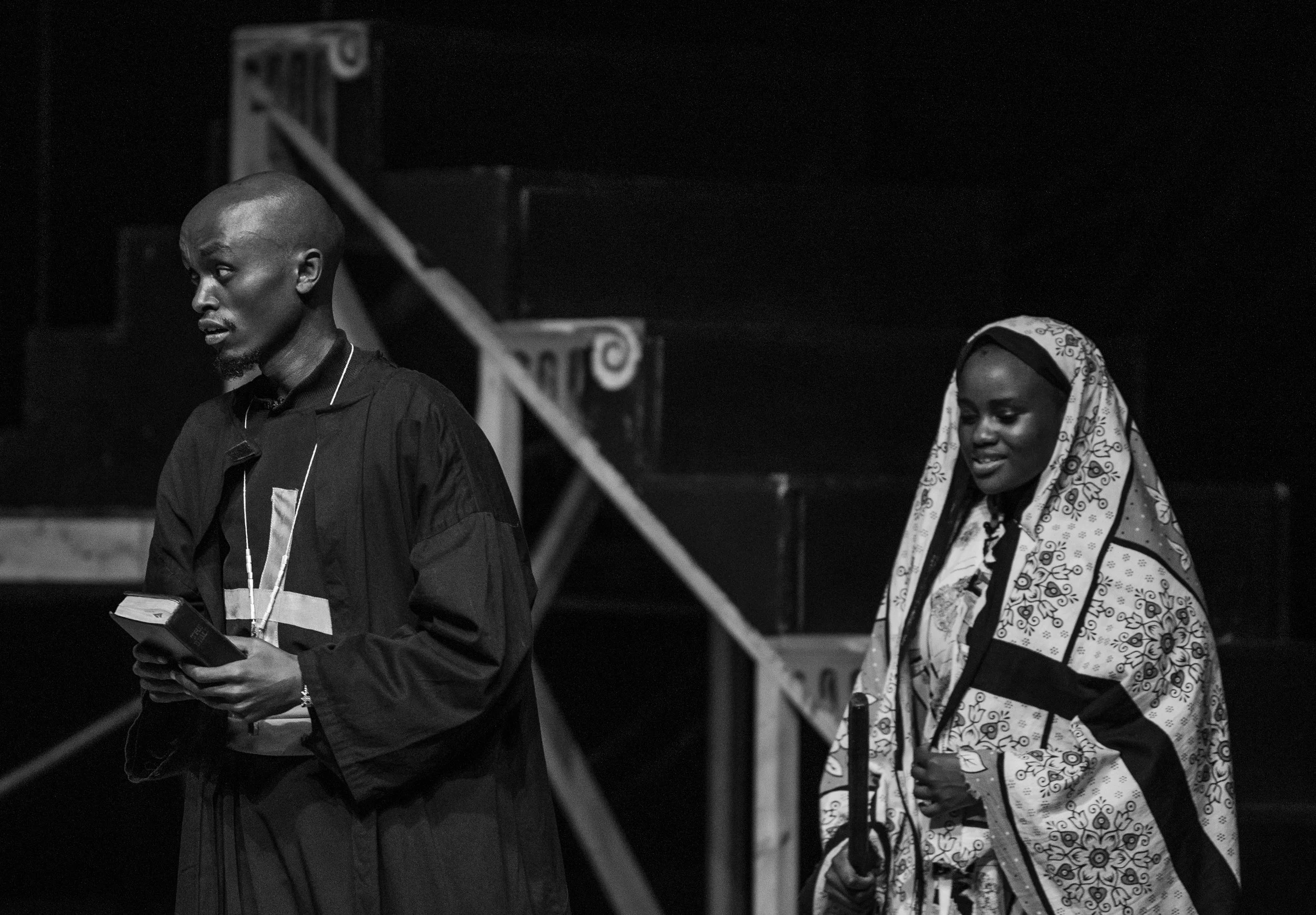 A black and white photo of a man and woman walking, the man holding a book, with a staircase in the background.