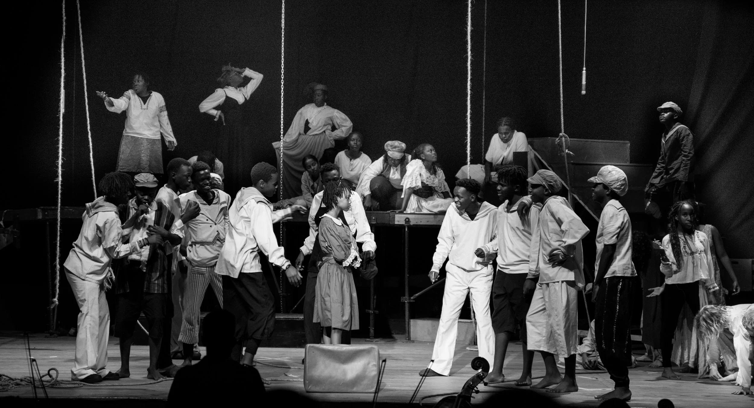 A black-and-white photo of a theatrical stage with actors performing. Several actors are standing and sitting, engaging in dialogue or action, with some in period costumes. The background features a set with hanging ropes and elevated platforms.