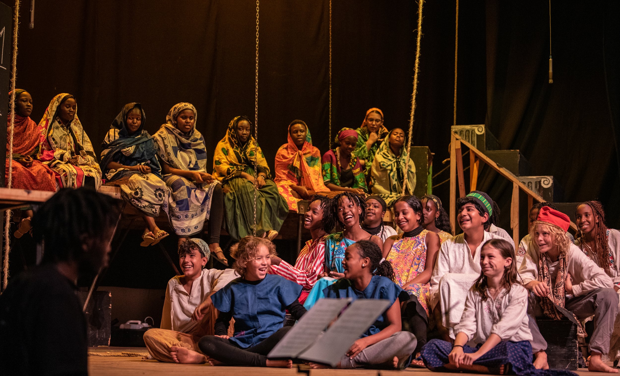 A diverse group of children and adults dressed in colorful costumes performing or rehearsing on a stage with a black backdrop, some sitting and some standing, sharing a joyful moment.