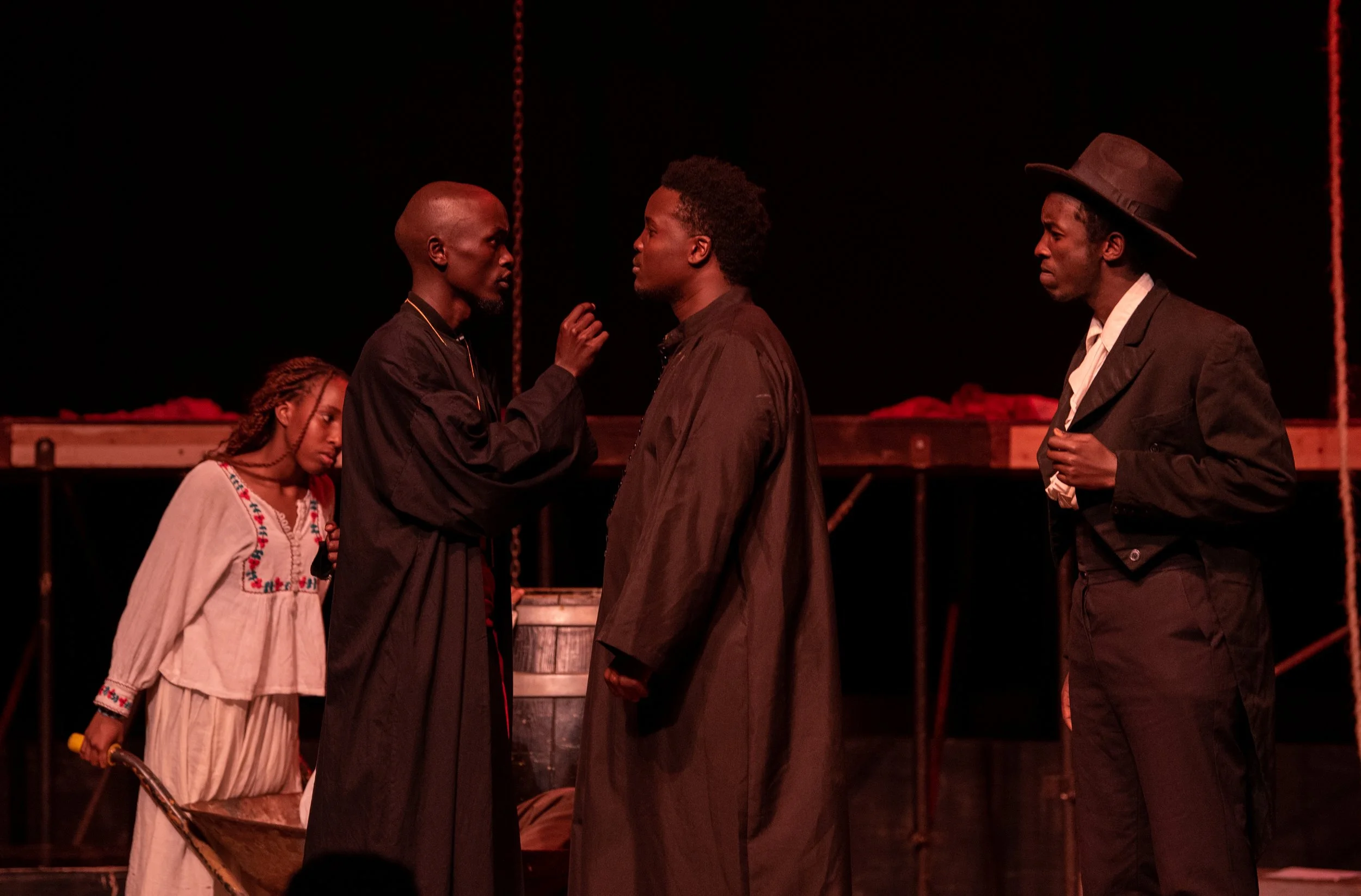 Four actors on stage in a theatrical performance, one woman and three men, dark background, dramatic scene.