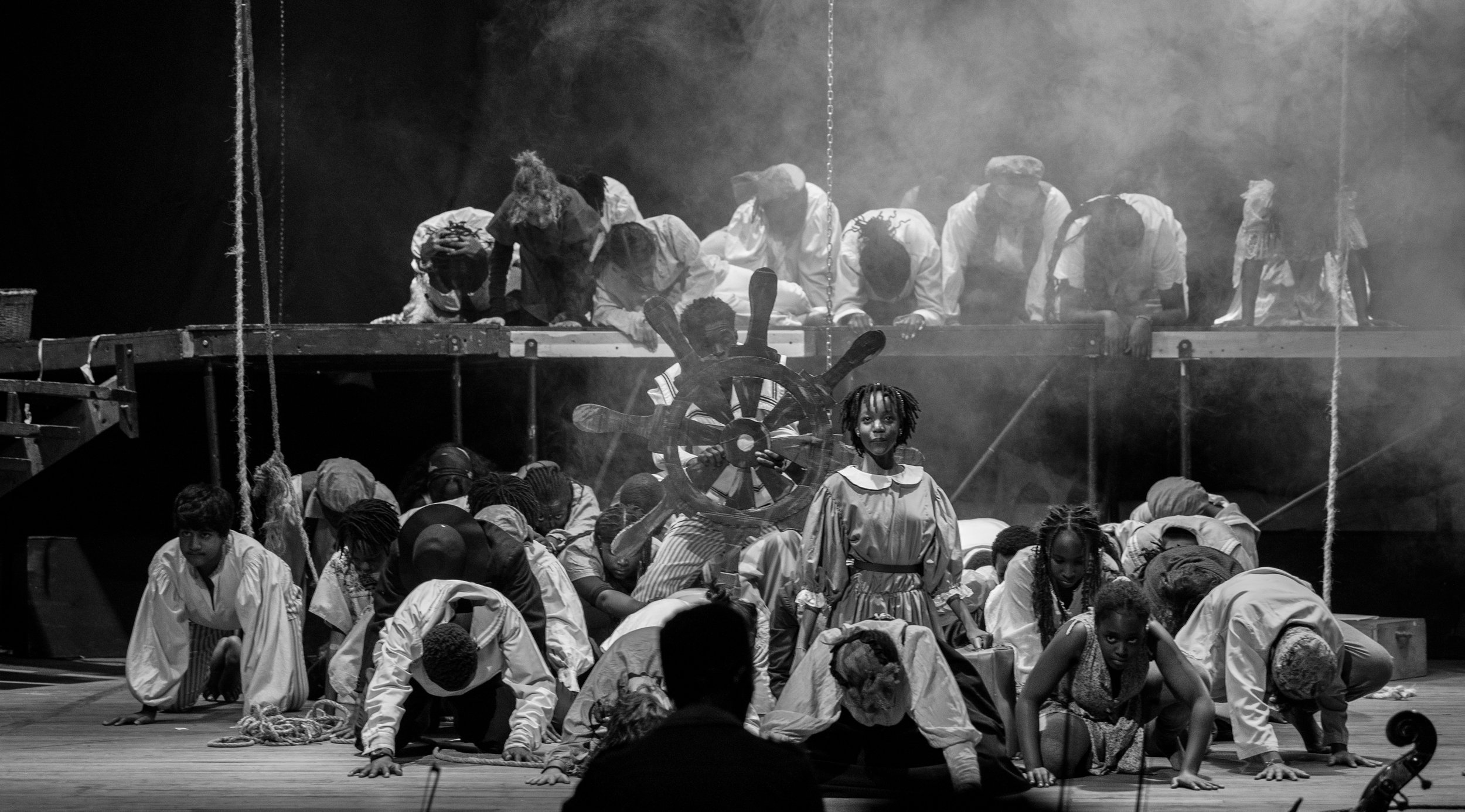 A theatrical stage performance with children dressed in period costumes, some kneeling or crawling, and others lying or standing, with a director or conductor in the foreground. The scene is set with fog effects, ropes, and a pirate ship steering whe