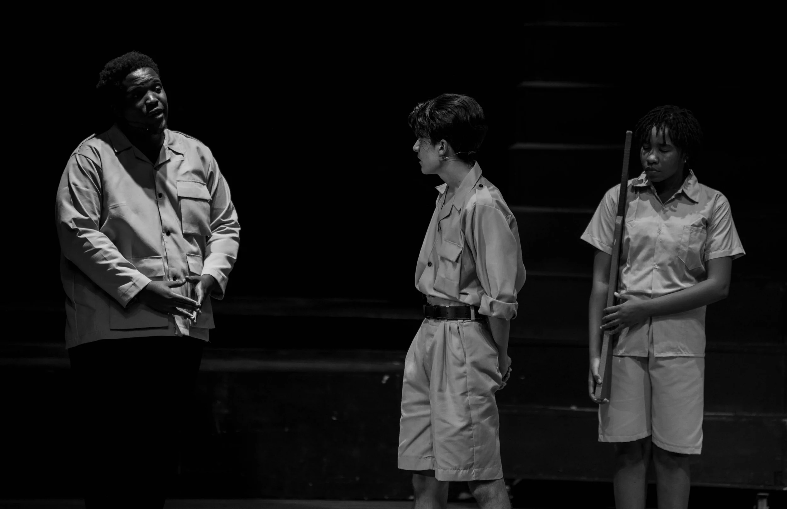 Three individuals engaged in a theatrical performance on stage, with the woman on the left speaking to the two young people on the right. The setting is dark with minimal lighting highlighting the actors.