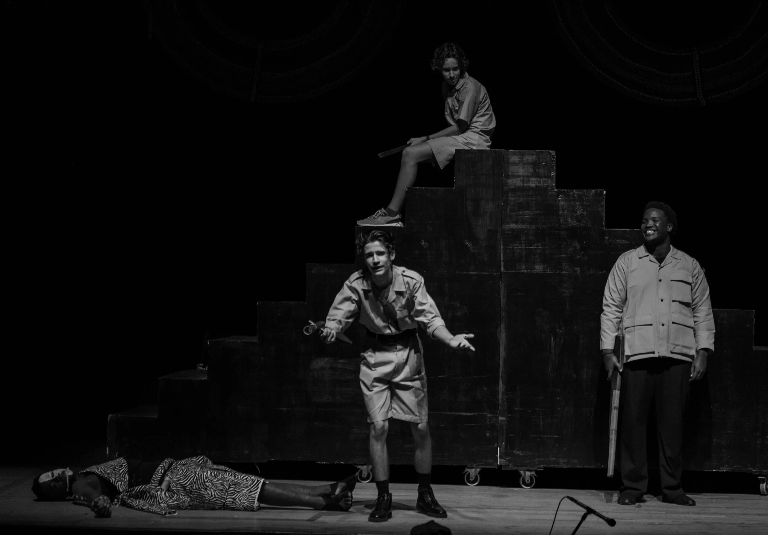 Black and white photo of four young actors on a stage with a dark background. One girl is sitting on a wooden block at the top, one boy is standing below her, another boy is lying on the floor, and a third boy is standing with a tool in his hand.