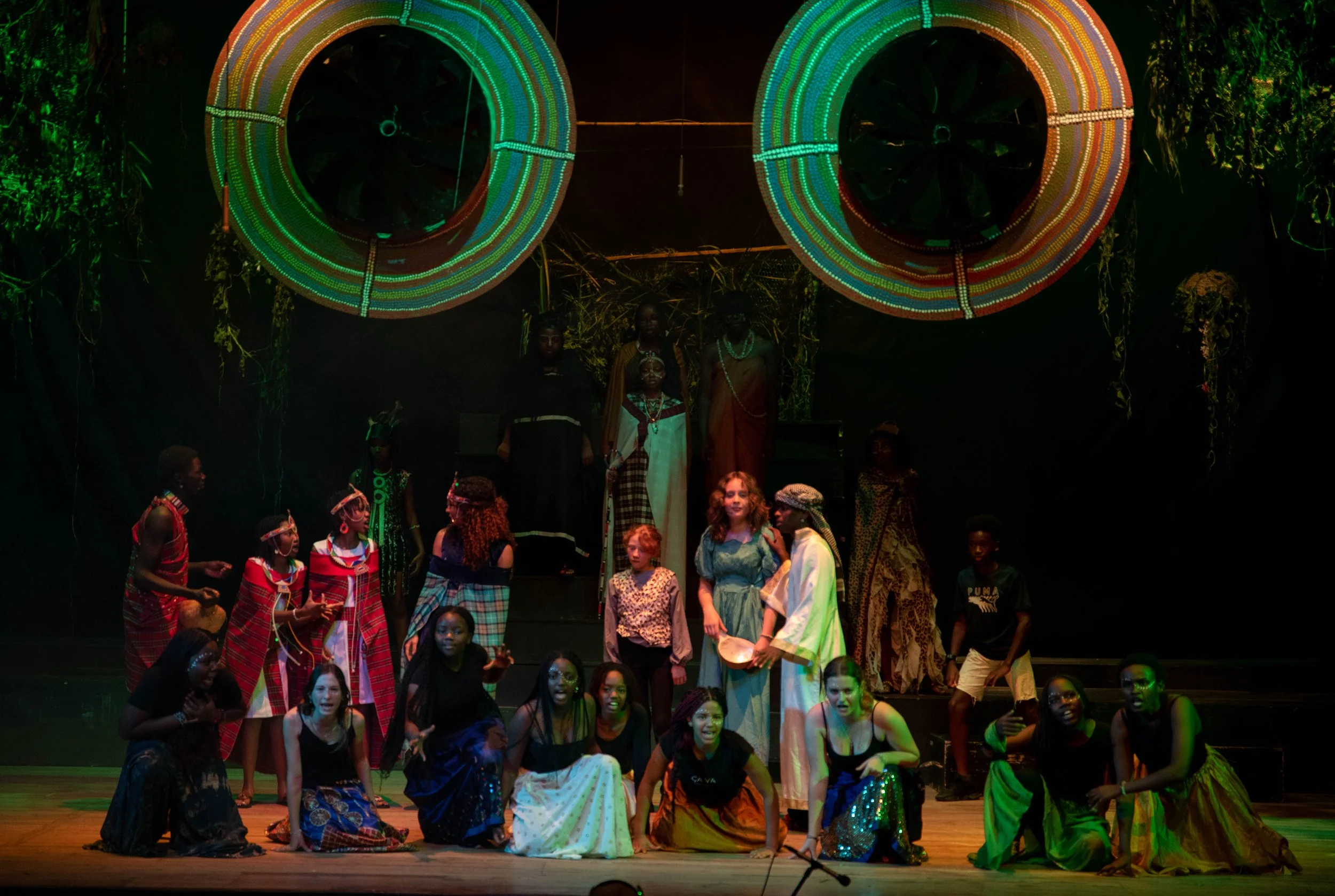 A theatrical stage performance featuring a diverse group of children and adults in colorful costumes. They are standing and kneeling in front of a dark backdrop with vibrant circular light decorations overhead. The scene appears lively and expressive