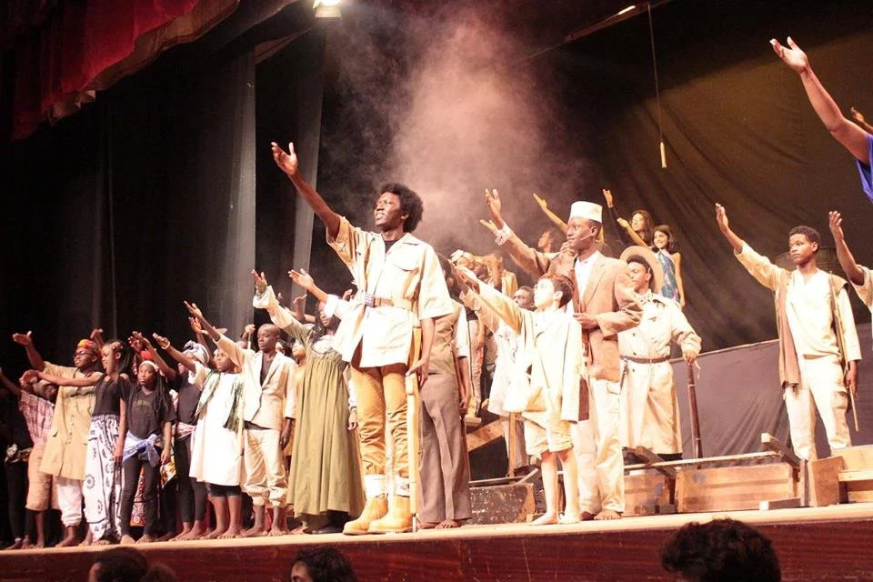 A group of performers on stage, raising their hands in a unified gesture, during a theatrical production. The stage is lit with warm lighting, and some performers are dressed in vintage or period costumes.