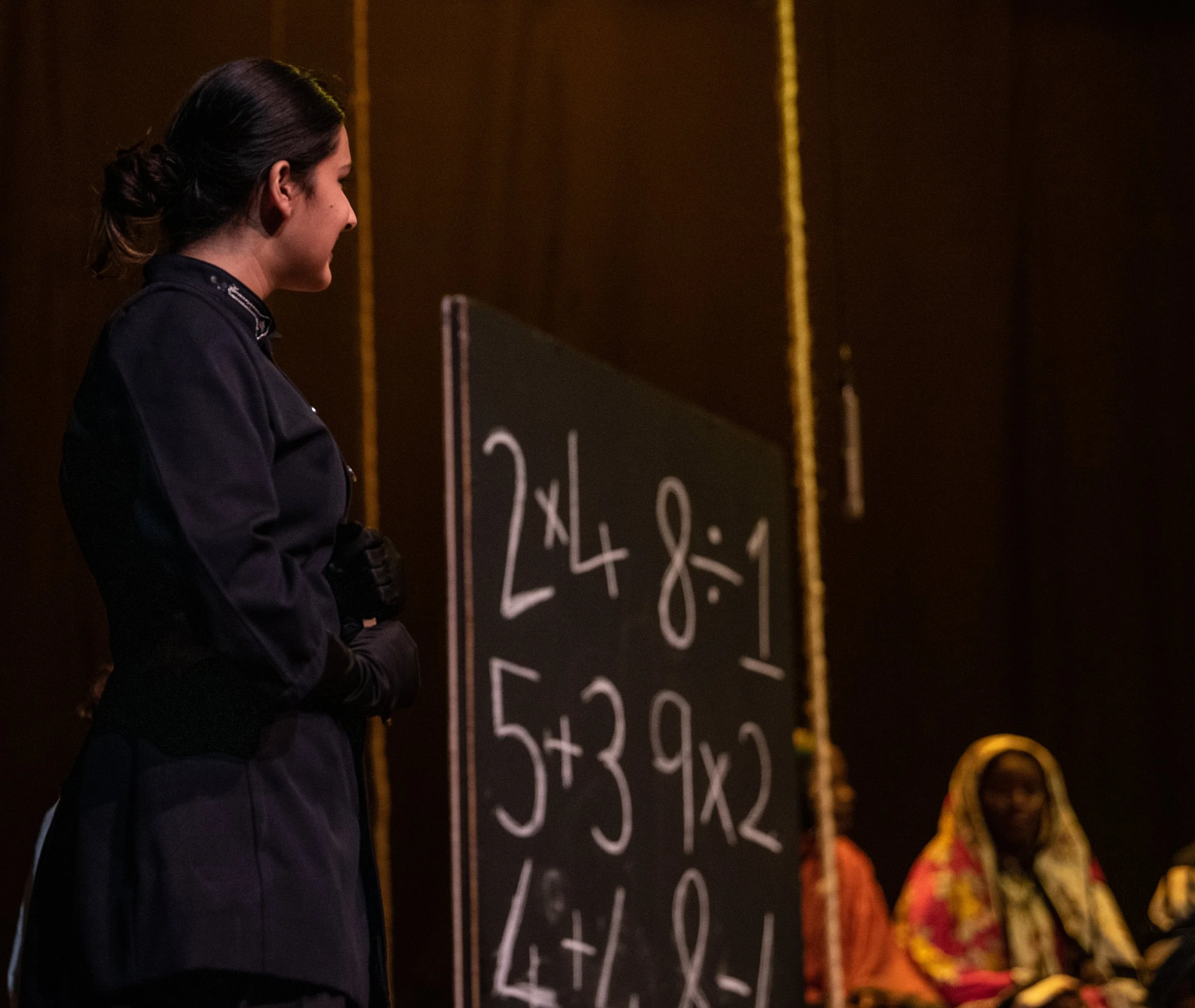 A woman dressed in black viewing a chalkboard with basic math problems written on it, and two women sitting nearby in colorful traditional clothing.