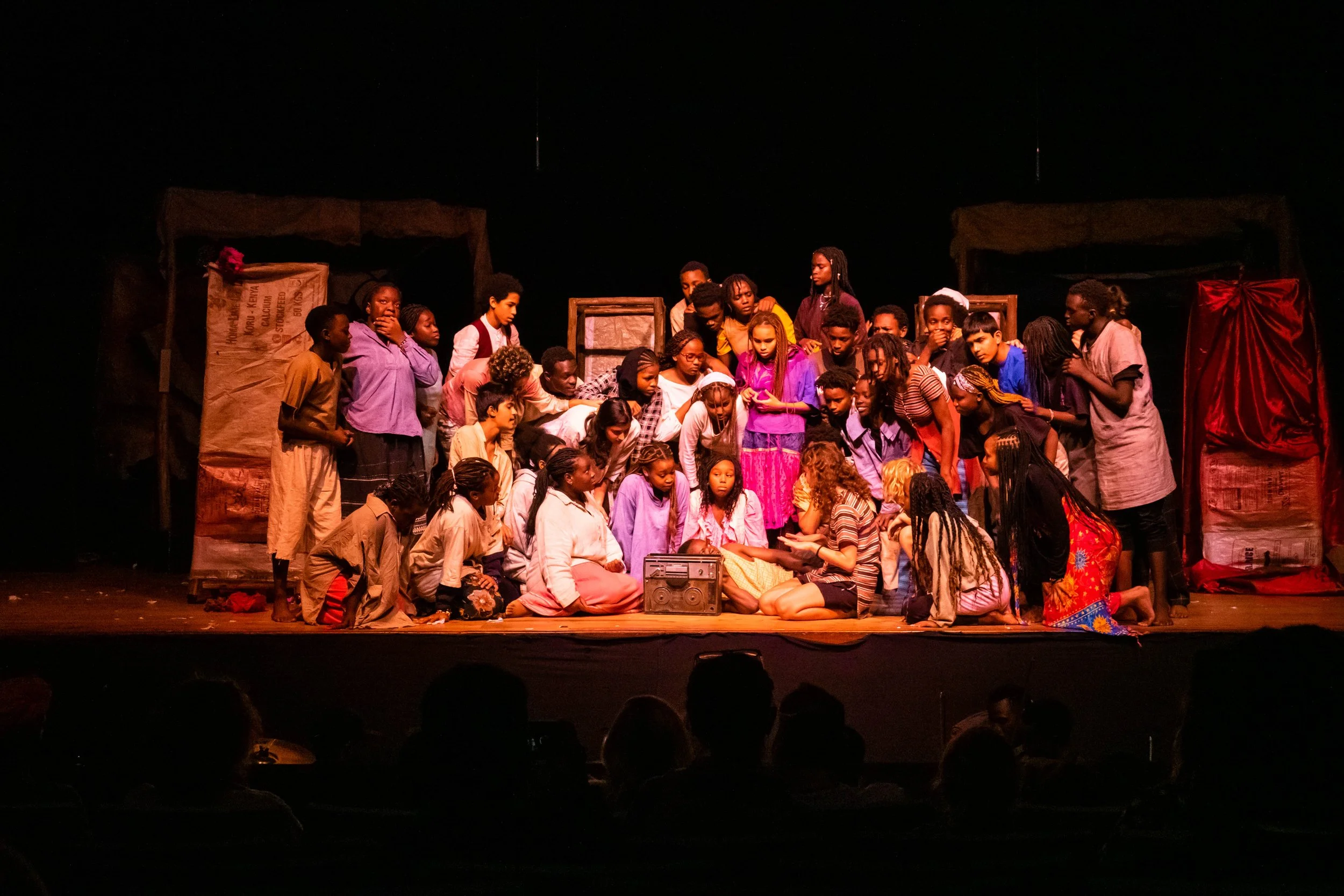 Scene from a theatrical play with a large group of children and teenagers on stage, some kneeling, some standing, in colorful costumes, scene set with makeshift structures, audience silhouettes visible in the foreground.
