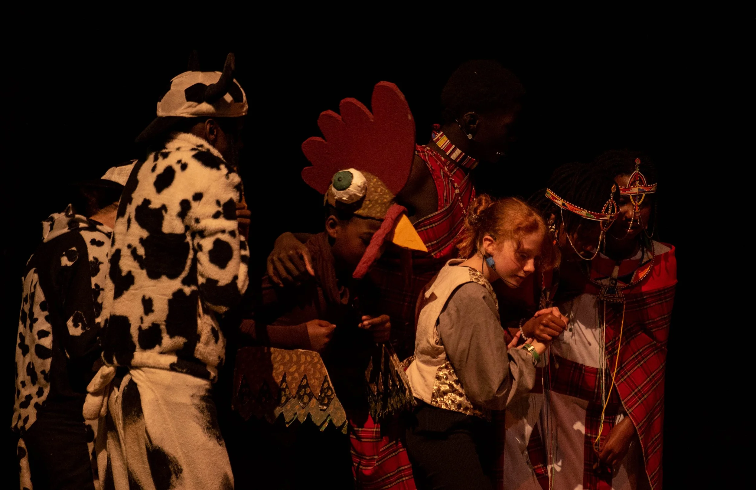Group of people dressed in traditional and cultural attire participating in a ceremony or performance, holding hands, with a dark background.