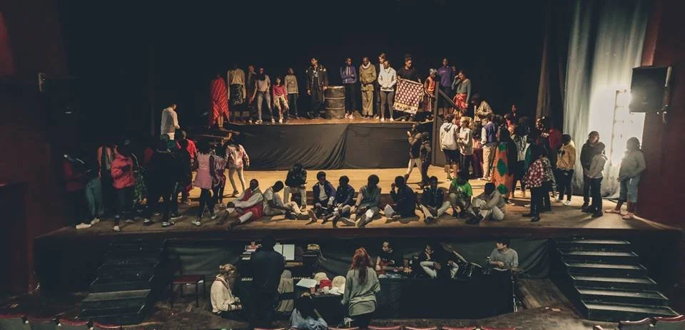 Group of children and adults on and in front of a stage in a theater, preparing for or practicing a performance, with some seated and others standing or walking.
