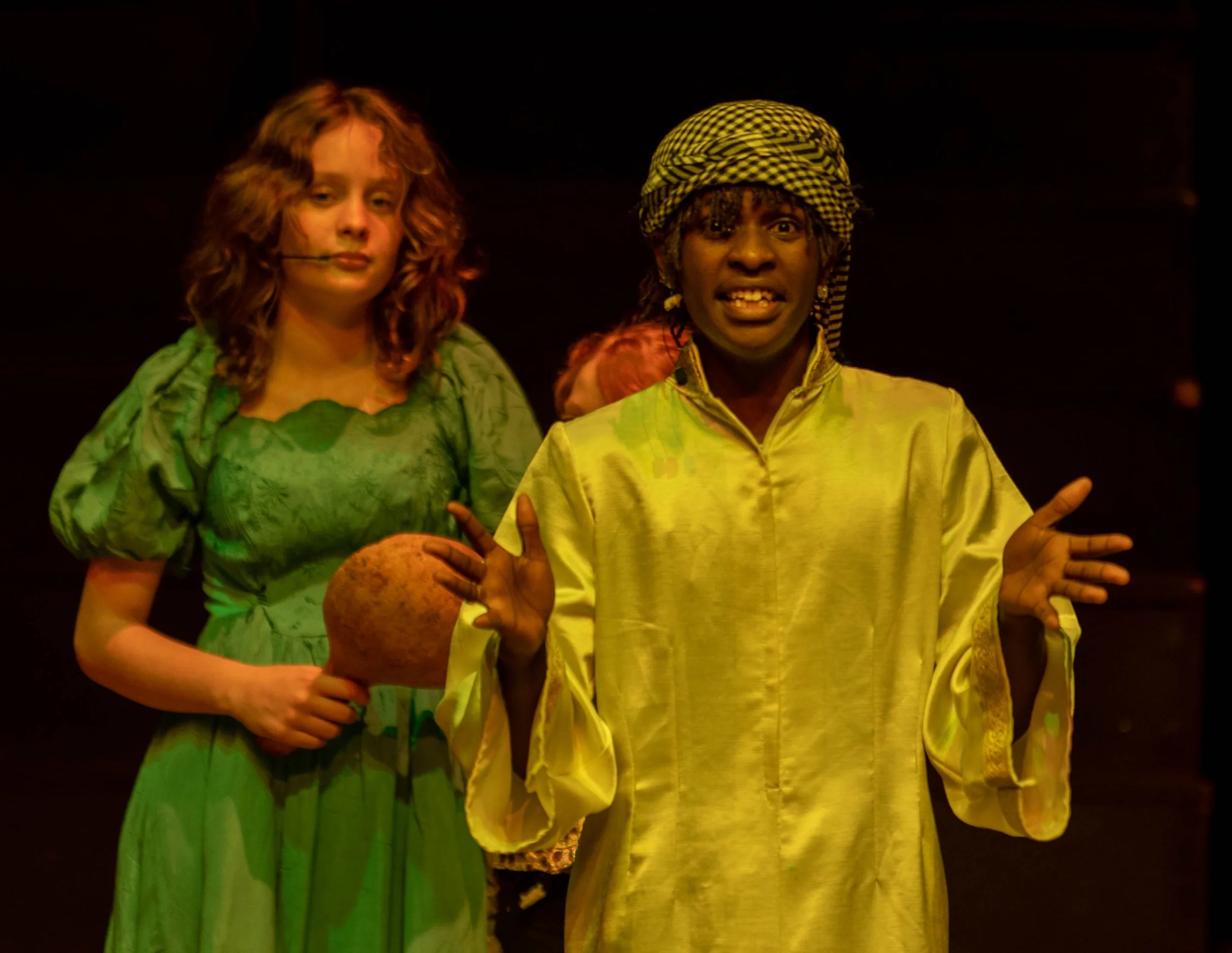 Two actors on stage, a woman in a green dress holding a large pomegranate and a man in yellow robes with a head wrap, gesturing with his hands and speaking.