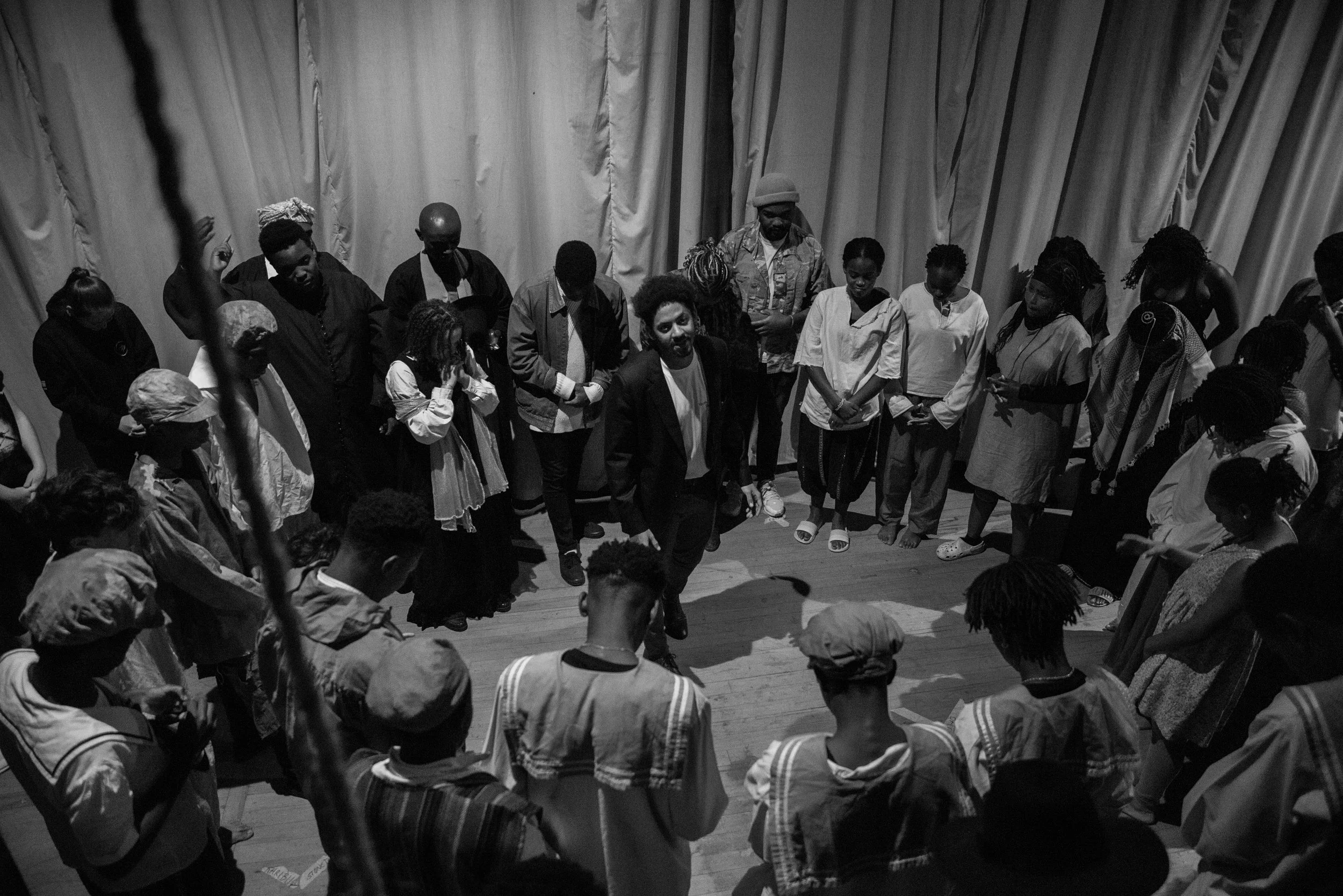 A black and white photo of a group of people standing in a circle on a stage, with one person dancing or performing in the center.