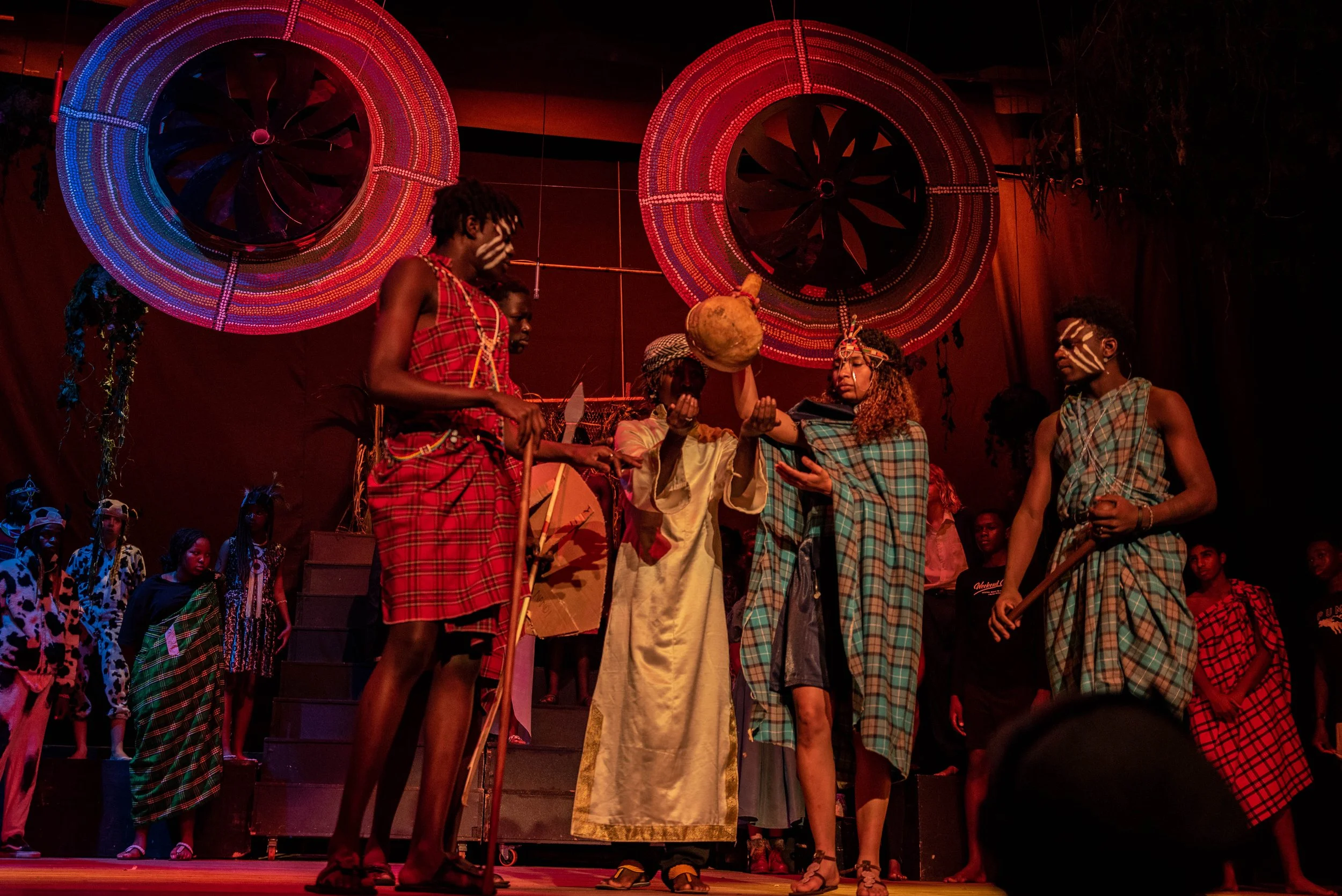 A group of performers in traditional tribal attire and face paint on stage, with large decorated circular backdrops above them.