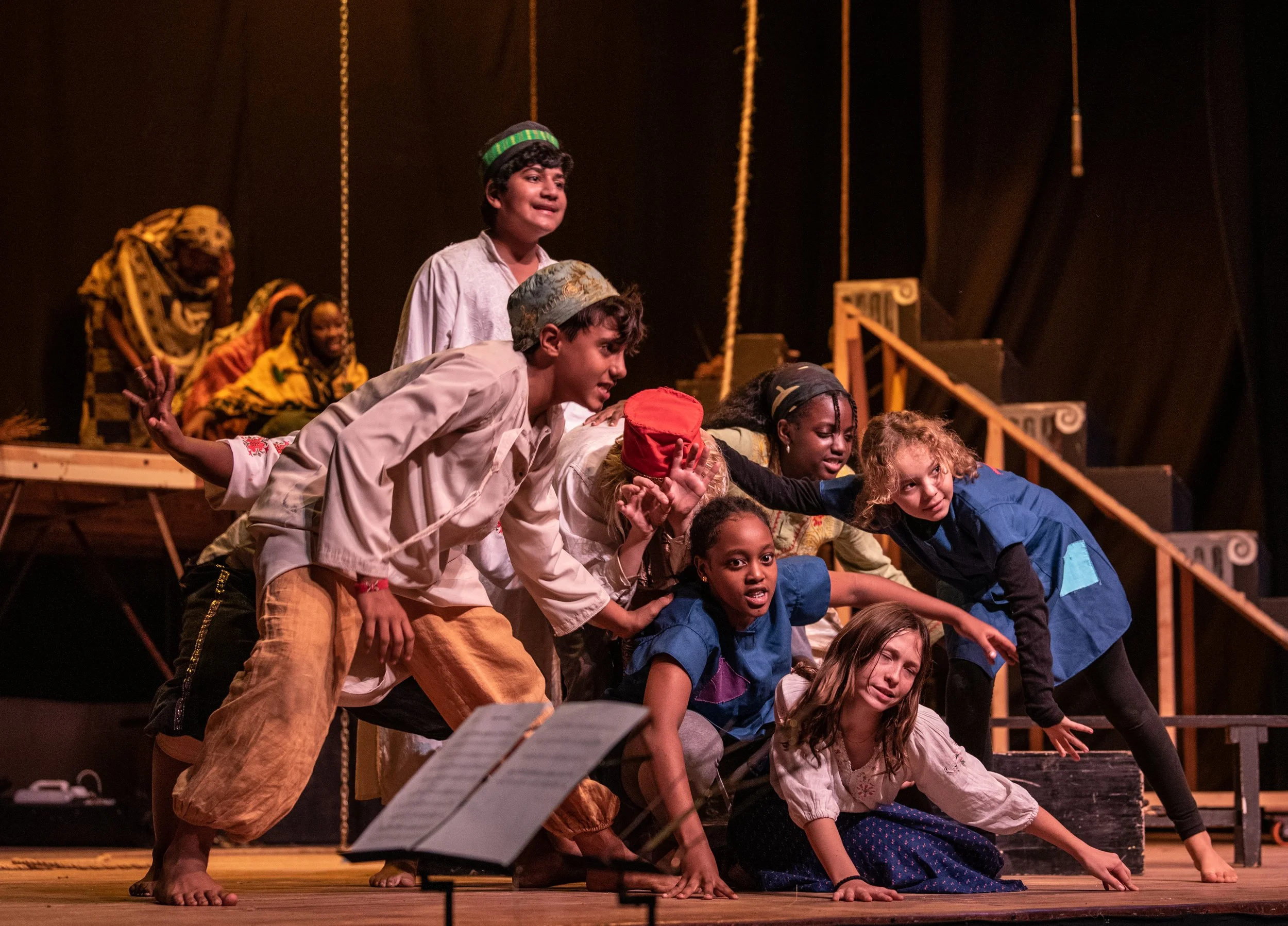Children performing in a theatrical play on stage, some standing and some crawling or kneeling, with a dark background and stage set including stairs and ropes.