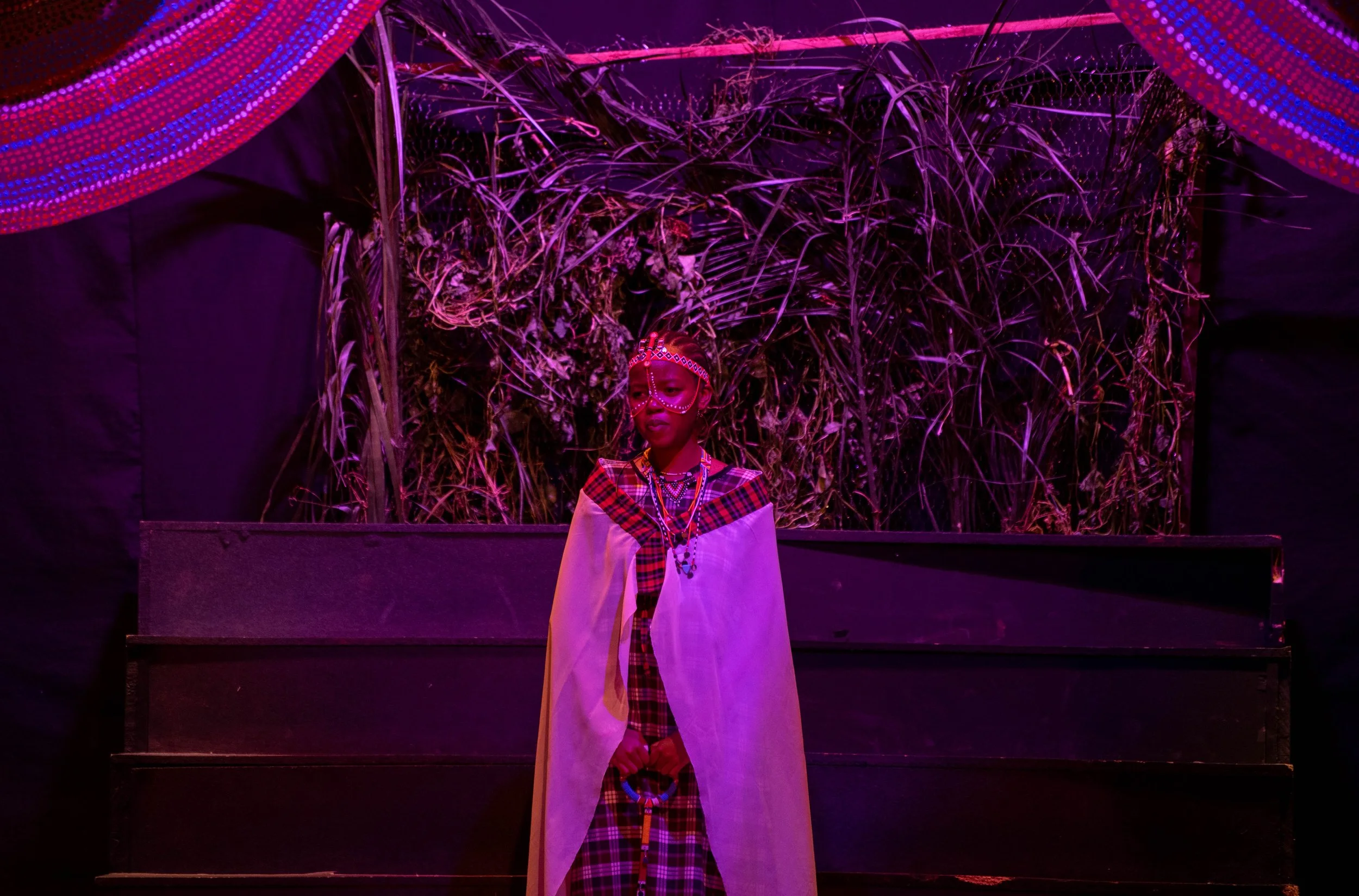 A person dressed in traditional tribal attire standing on a stage with a backdrop of natural elements, illuminated by purple lighting.