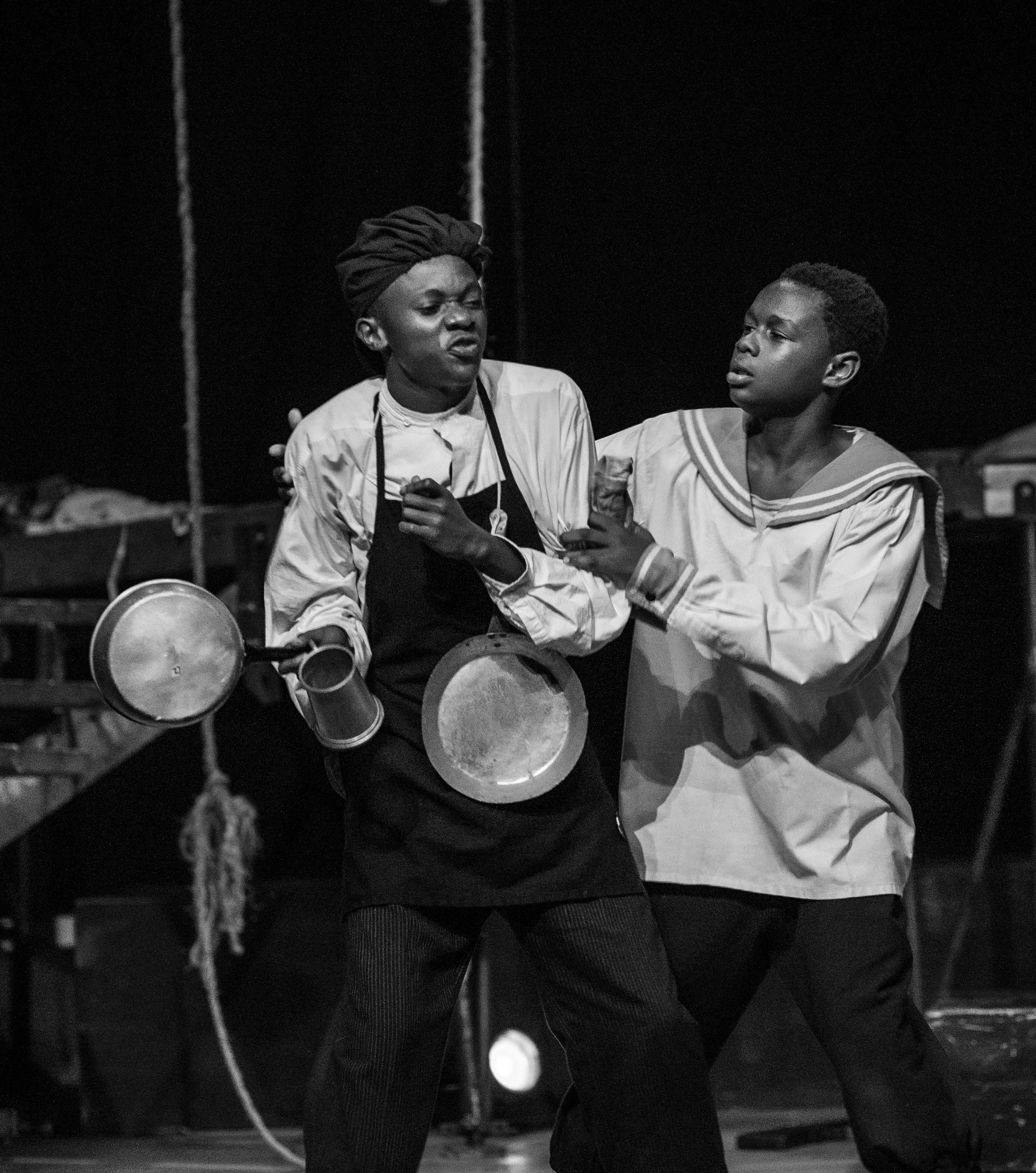 Two actors dressed in theatrical costumes performing on stage, one with a towel on his head and an apron, the other in sailor attire, engaged in a dramatic scene.