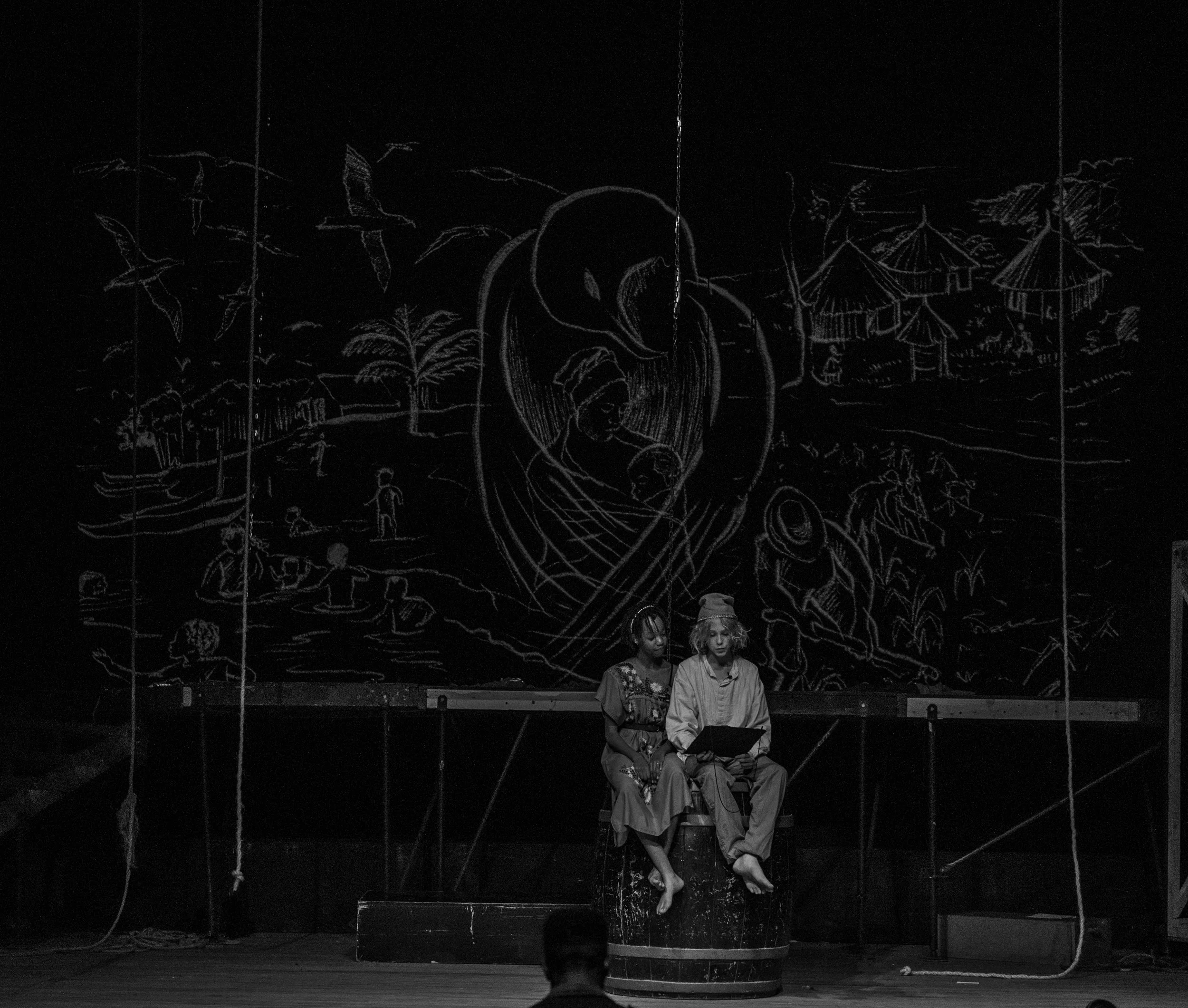Stage scene with two performers sitting in a barrel prop, a dark background with a chalk drawing of a village scene, people, and birds.