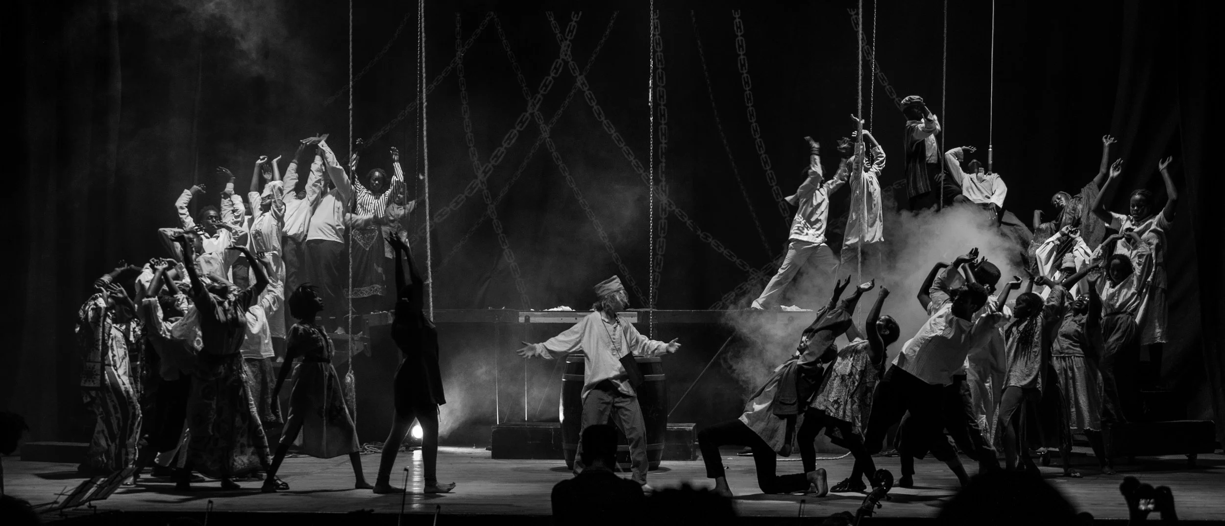 A theatrical stage performance with numerous actors in costume, some on the ground and others elevated, with dramatic lighting, smoke, and chains hanging from above.
