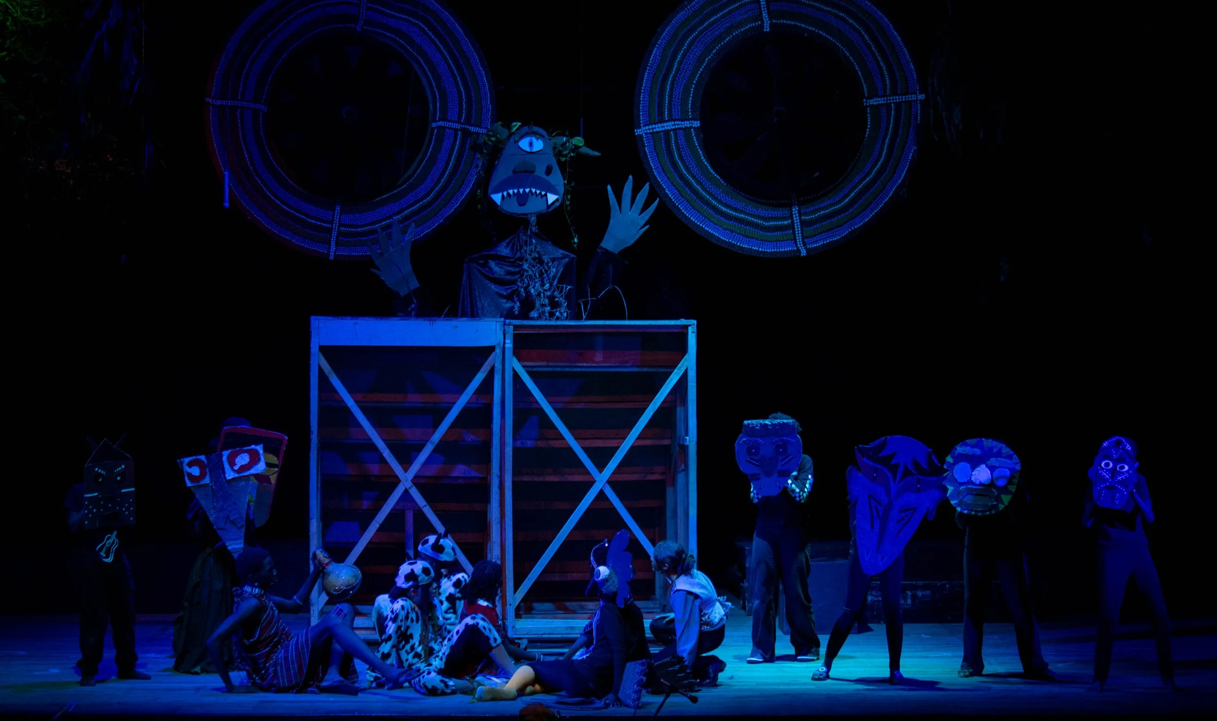 Children wearing dragon, owl, tiger, face masks on stage with puppet in costume, stage scene with illuminated circular props overhead.