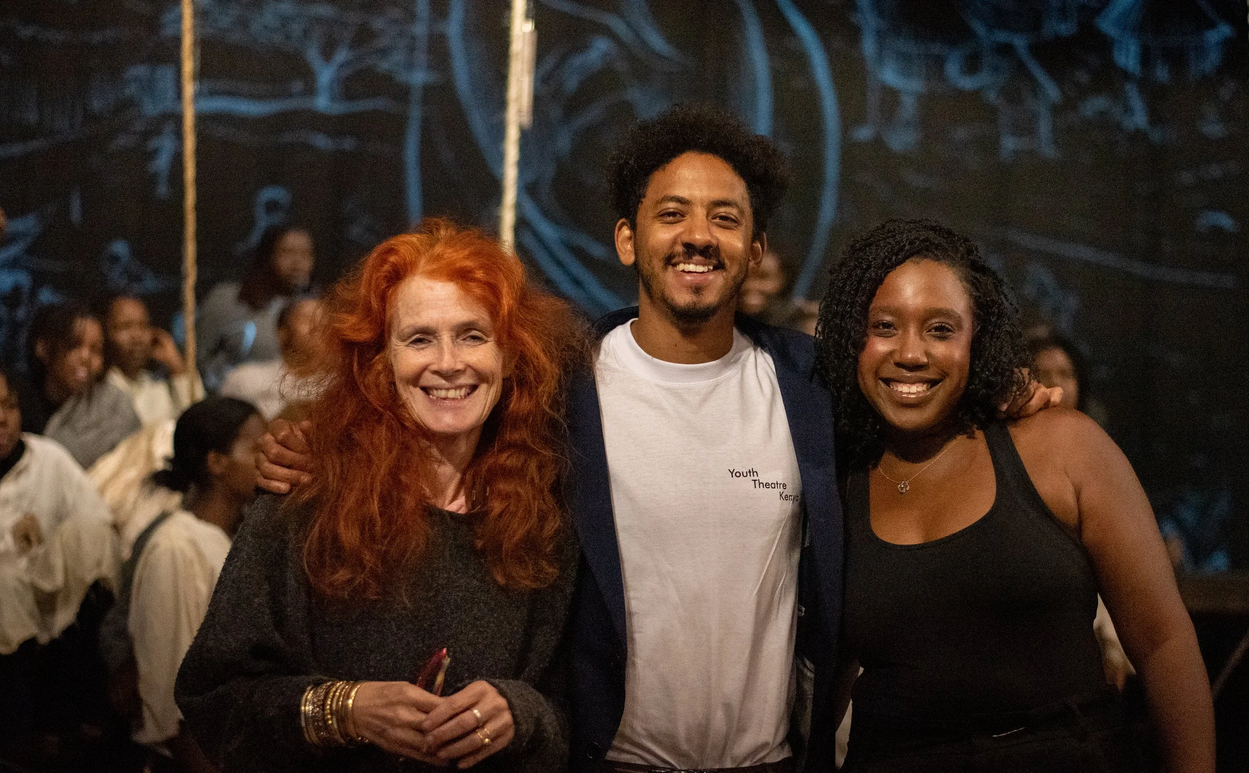 Three smiling people standing close together at an indoor event, with a group of people in the background, in front of graffiti-style artwork.