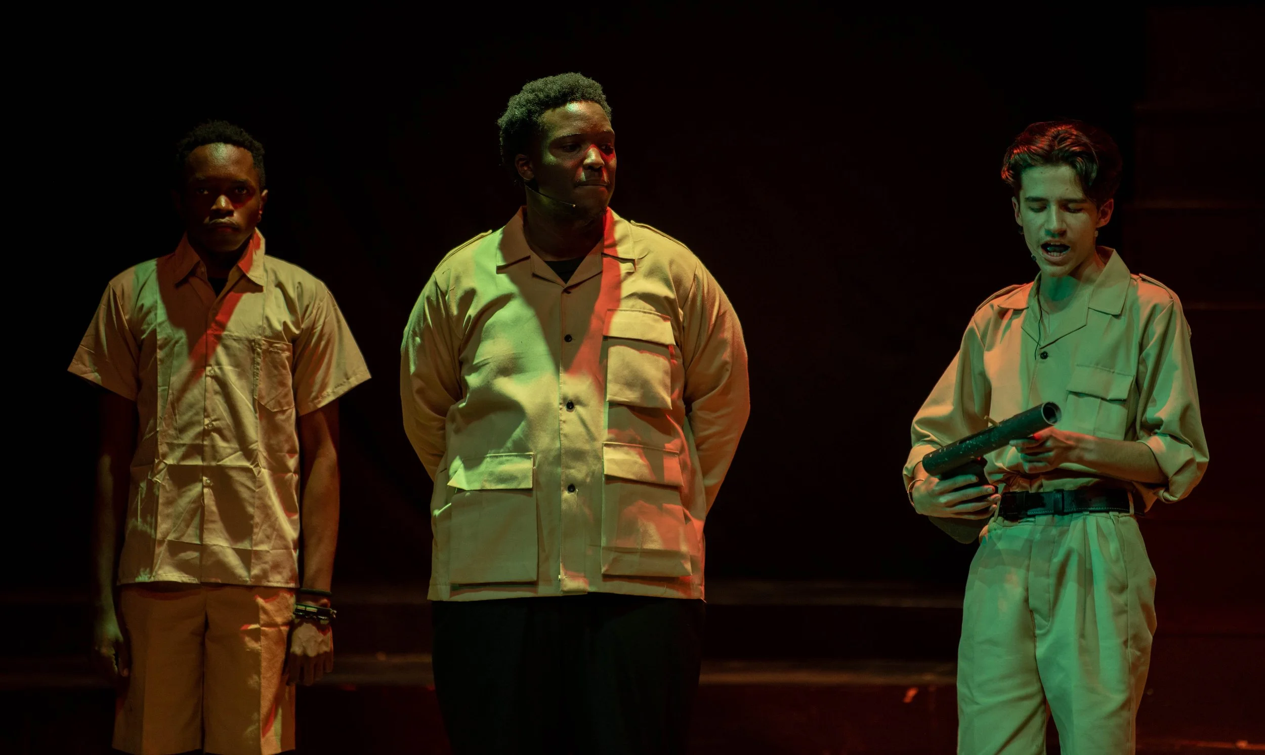 Three young men in tan uniforms standing on stage with dark background, one holding a pipe, during a theatrical performance.