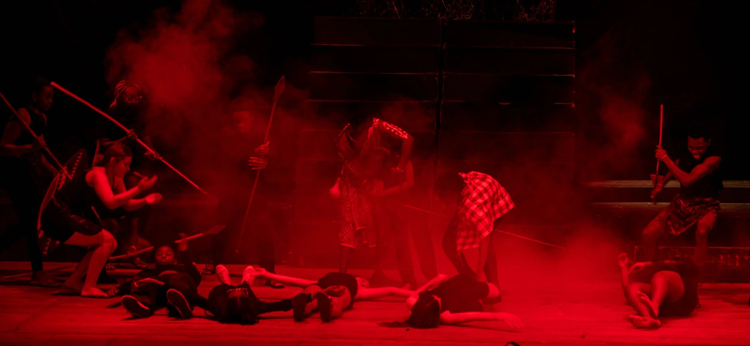 Children performing on stage with red lighting and smoke effects, some lying on the floor and others holding spears or other props.