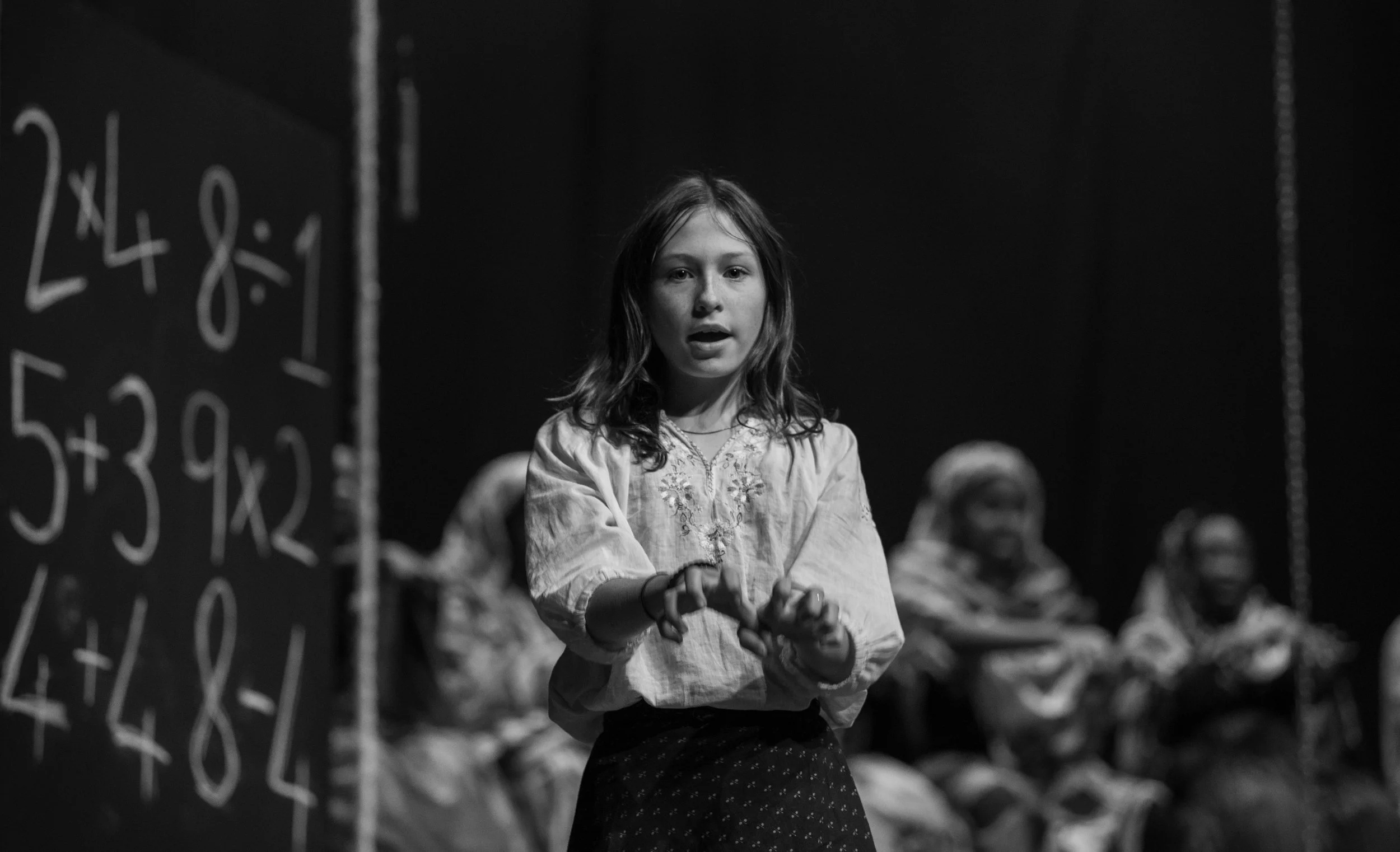 A young girl standing in front of a blackboard with math problems, with other children sitting in the background in a dark room.