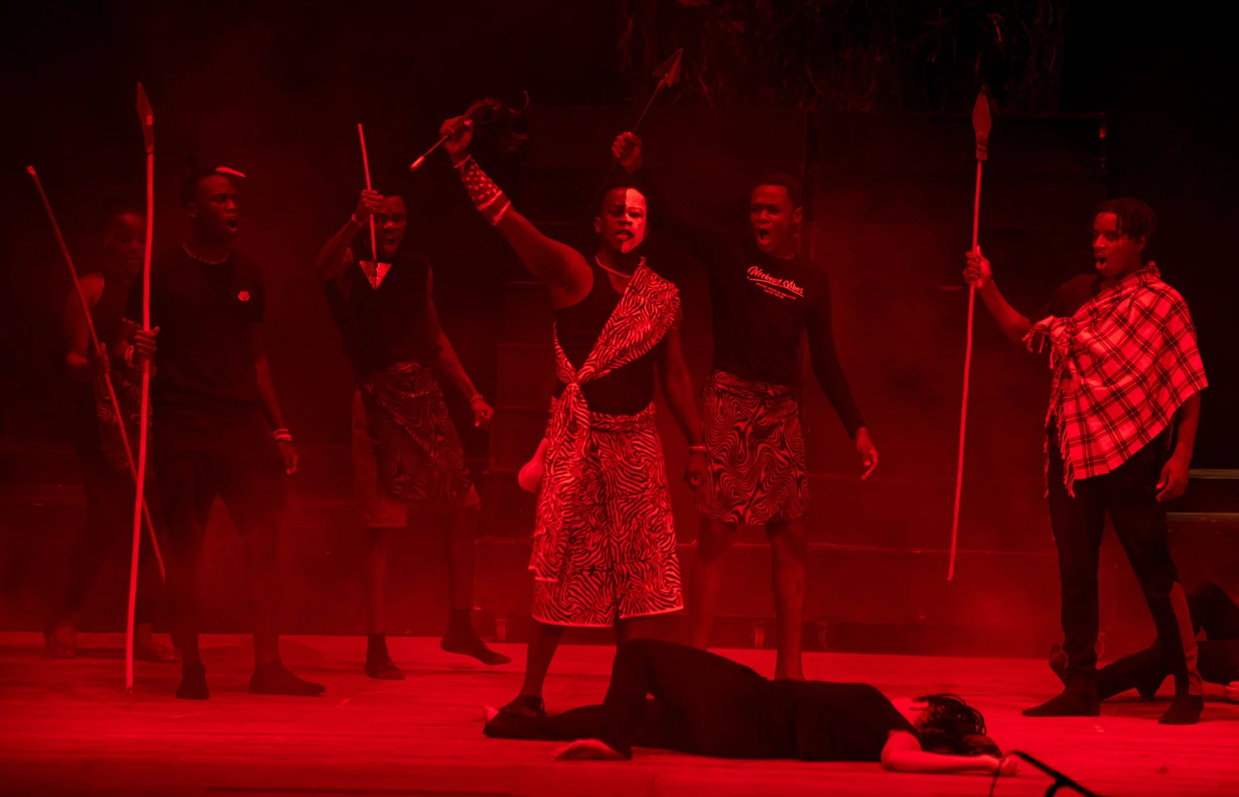 The image depicts a theatrical scene with several actors on stage, mainly dressed in black and wearing traditional African-inspired clothing with bold patterns. One actor stands center stage, holding a spear, with others surrounding and pointing spea