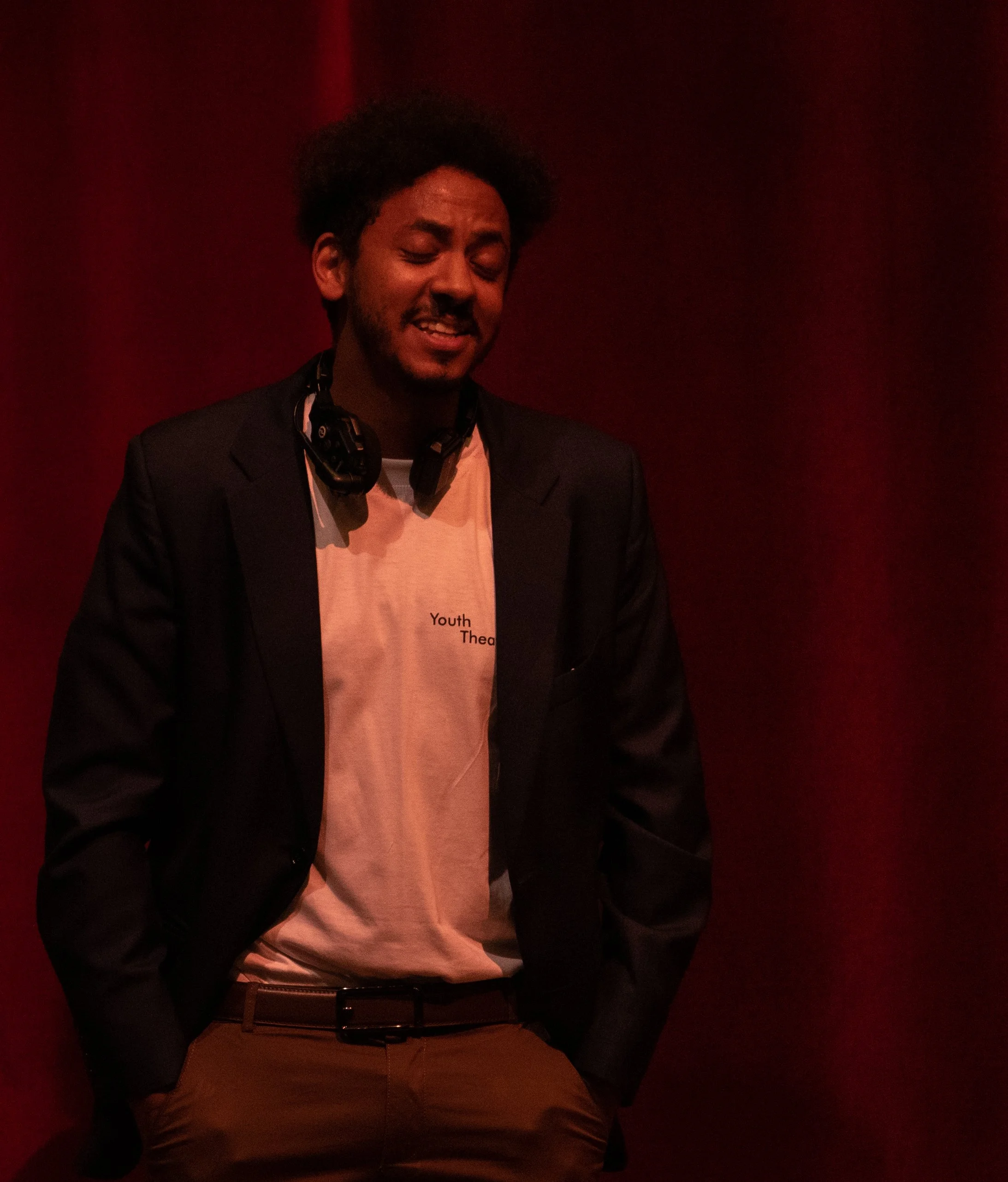 A man with curly hair and a beard wearing a black blazer, beige T-shirt, and brown pants stands against a dark red background with his hands in his pockets, smiling with his eyes closed.