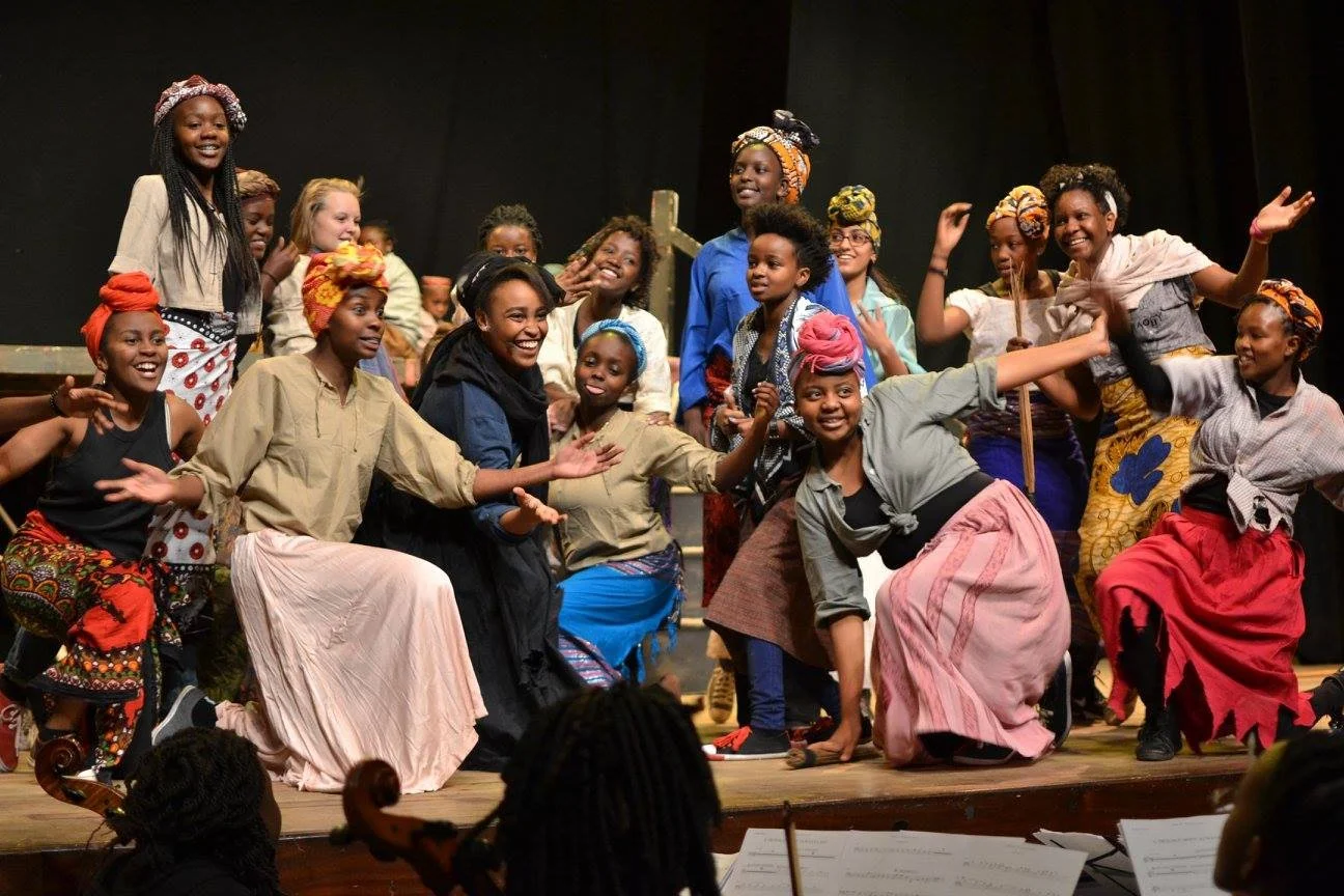 Group of diverse women and girls on stage dressed in colorful traditional African attire, smiling and dancing.