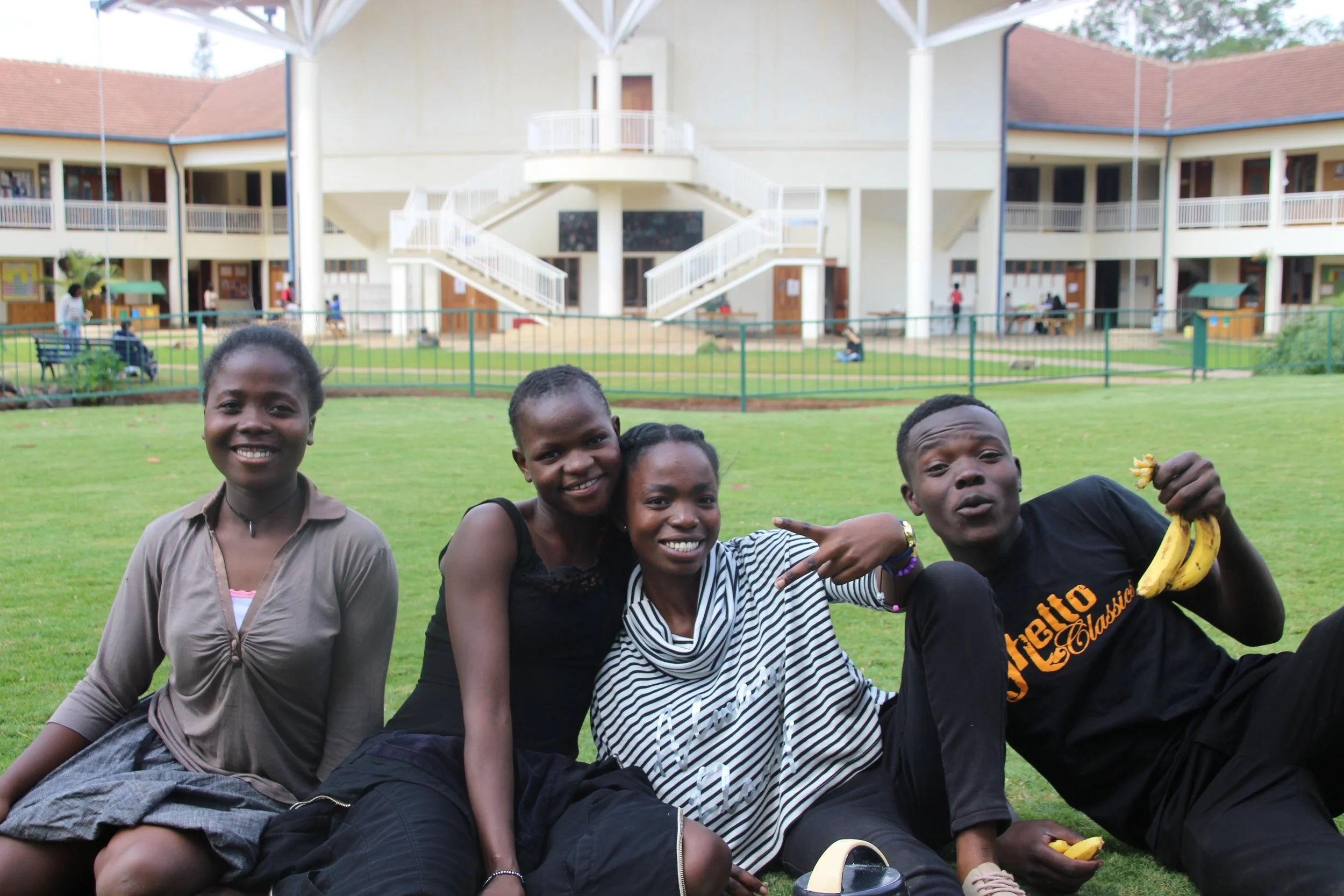 Four young people sitting on grass in front of a school building, smiling and posing for the photo, with bananas and other snacks.