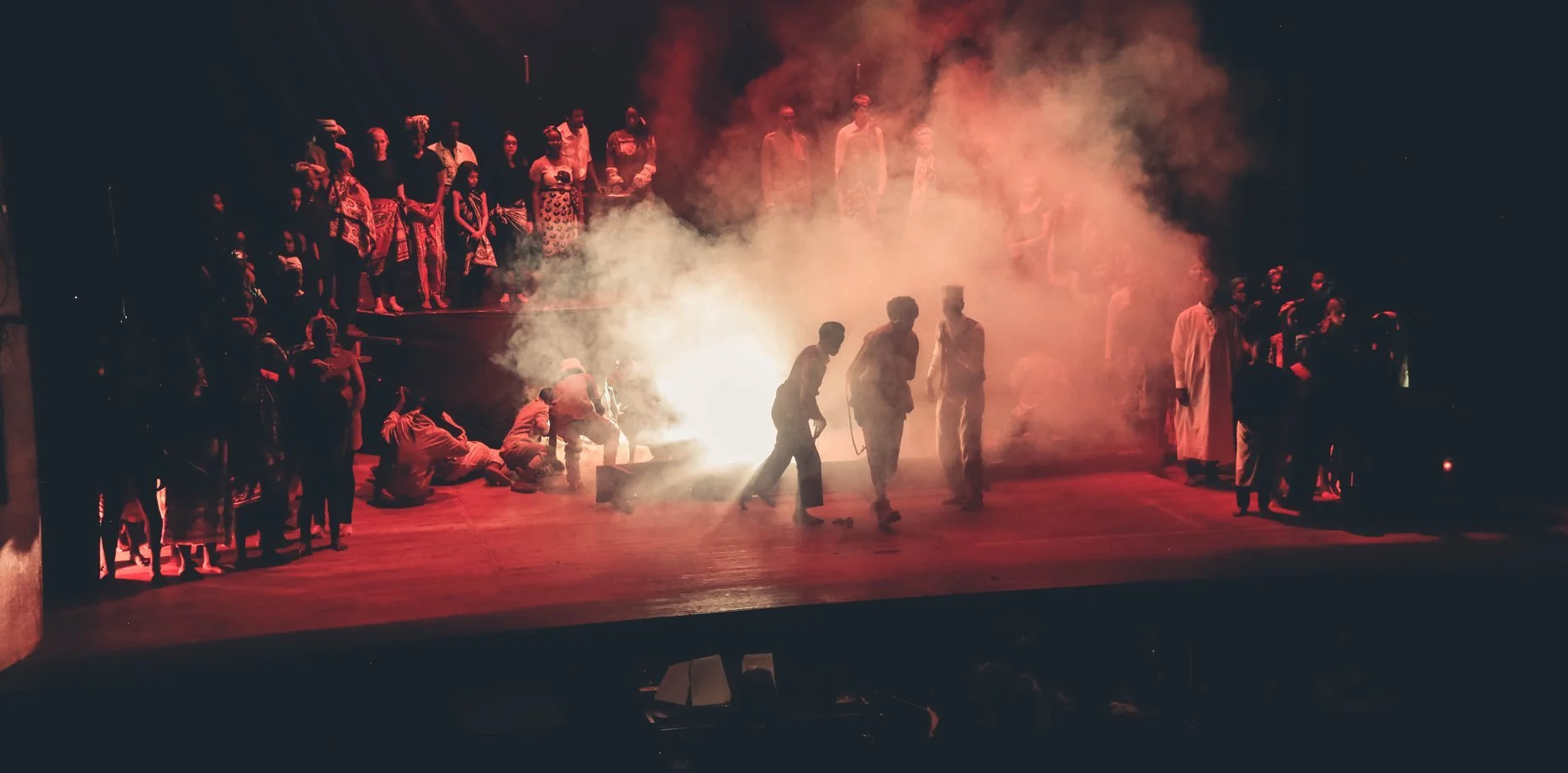 Stage performance with a group of people, some gathered around a fire with smoke, and others watching from the background, all dressed in traditional and modern clothing.