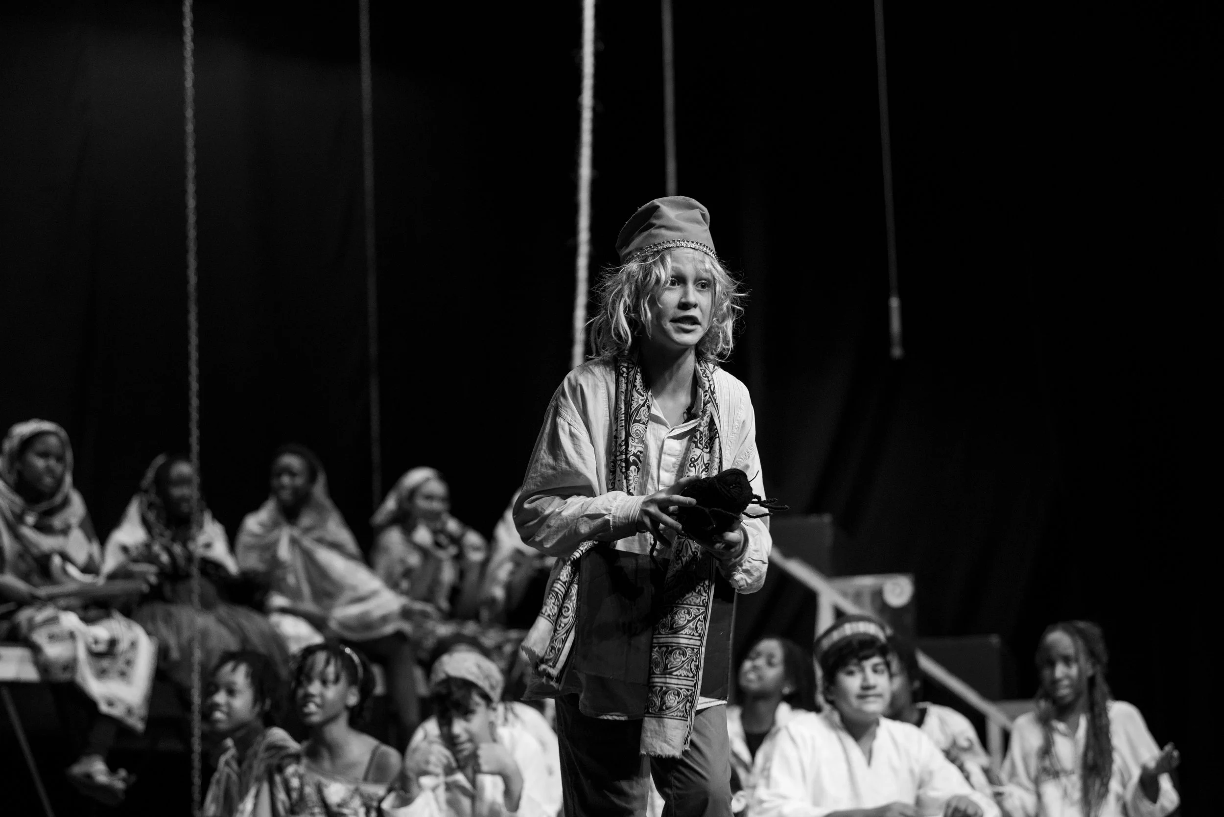 A woman with curly hair and light-colored clothing appears to be performing on stage, holding a prop, with a group of children and women sitting and watching in the background.