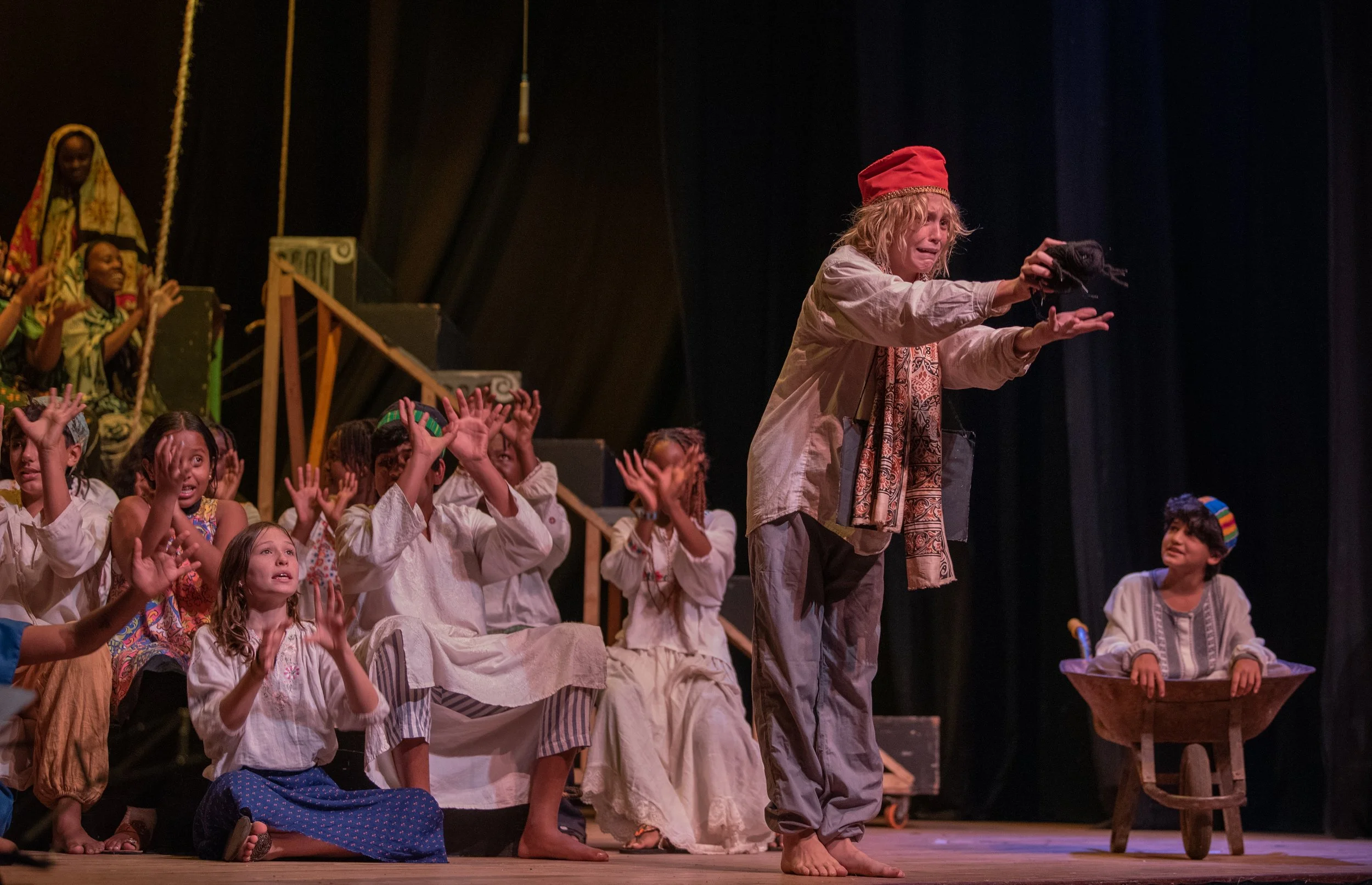 A theatrical stage scene with children and a woman dressed in medieval-style costumes, some children on the left raising their hands, the woman holding a black object, and a boy in a wheelbarrow on the right, all in front of dark curtains.