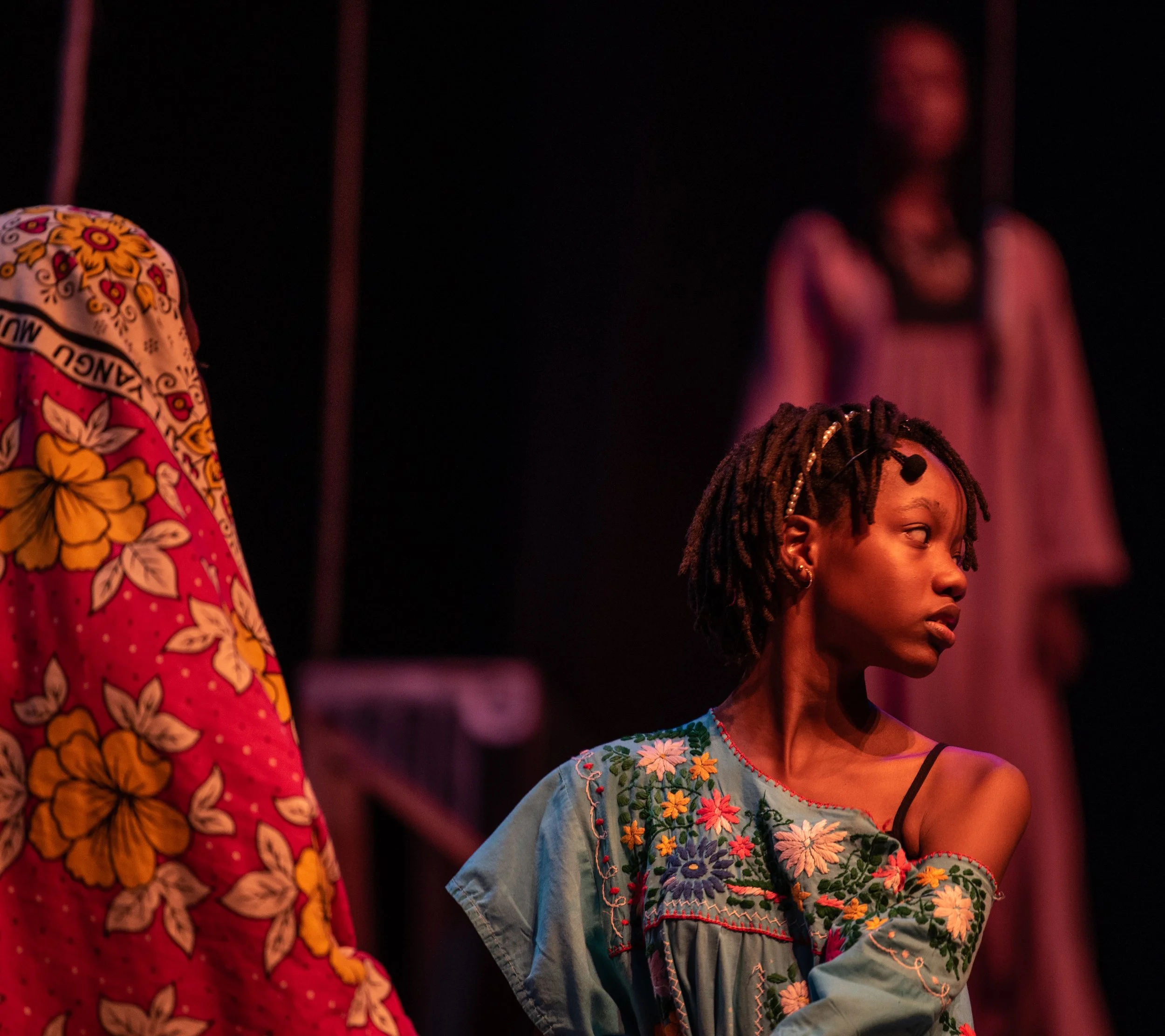 A young woman with braided hair and earrings looking to her right, wearing an embroidered blue dress with floral patterns, on stage with another woman in colorful clothing in the background.