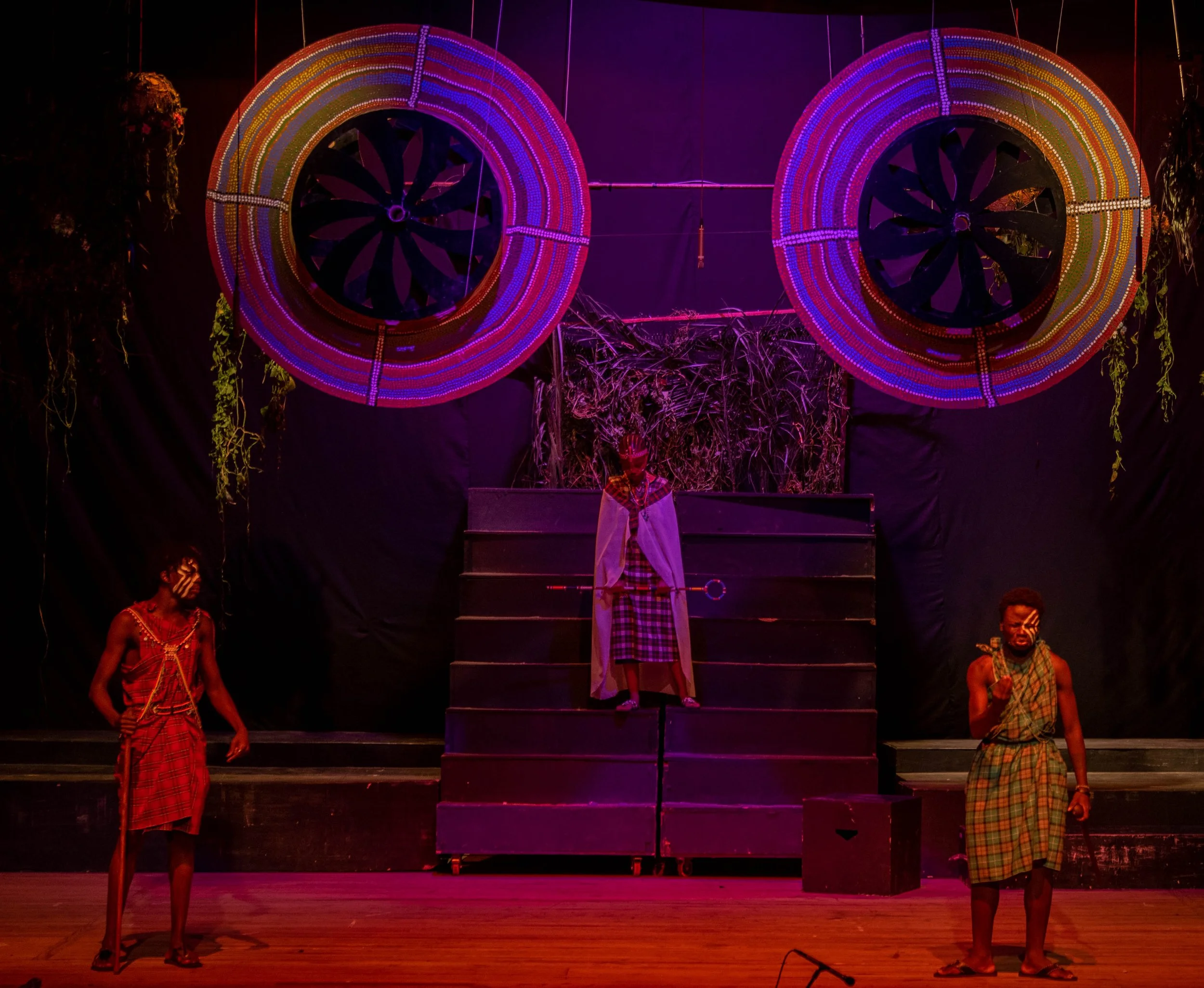 Theatrical stage with three actors in traditional Maasai attire, one standing on the staircase and two at stage level, with large colorful circular decorations overhead and a backdrop of foliage, illuminated by vibrant purple and pink lighting.