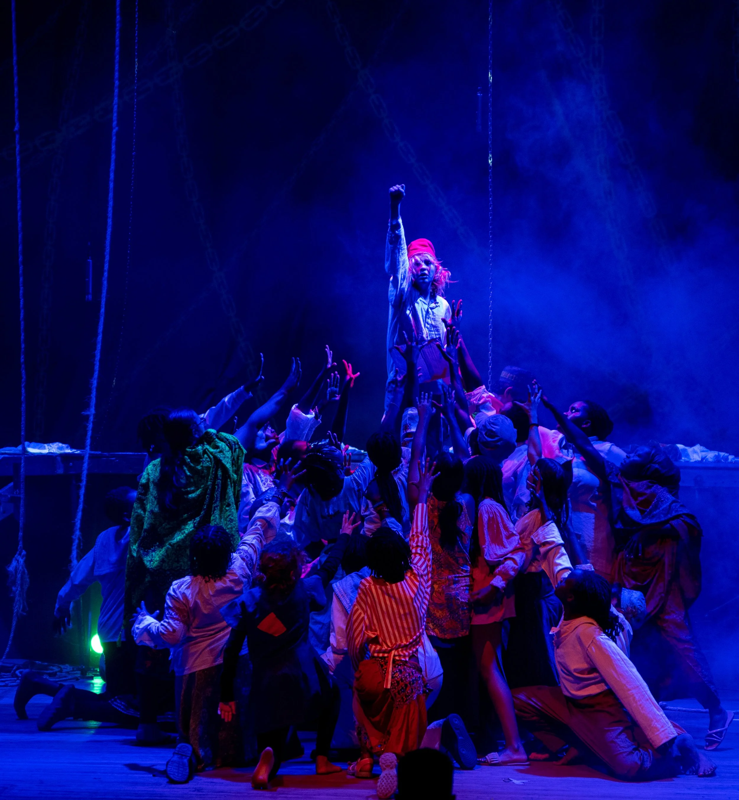 A theatrical performance featuring a person standing on an elevated platform, raising a fist, surrounded by a group of children reaching towards them. The scene is illuminated with blue stage lighting and fog.