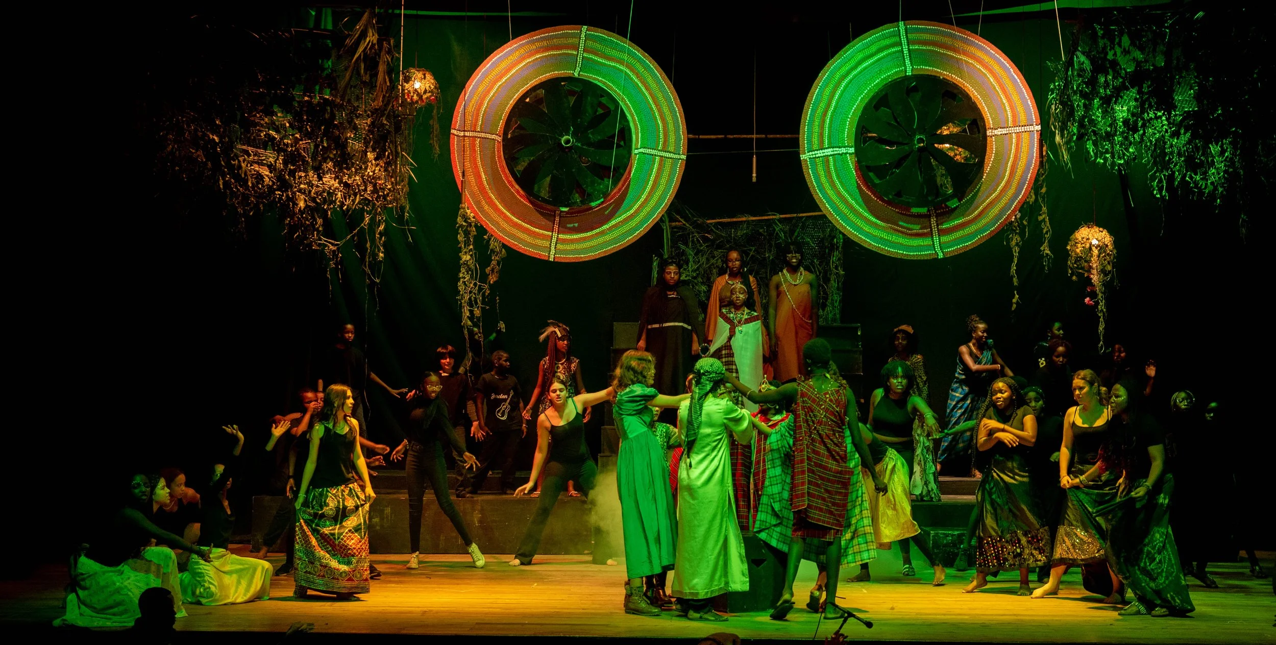 The stage features a diverse group of performers, including children and adults, engaging in a theatrical or cultural performance. The background includes large, colorful, circular light installations, with greenery and flowers hanging from above, cr