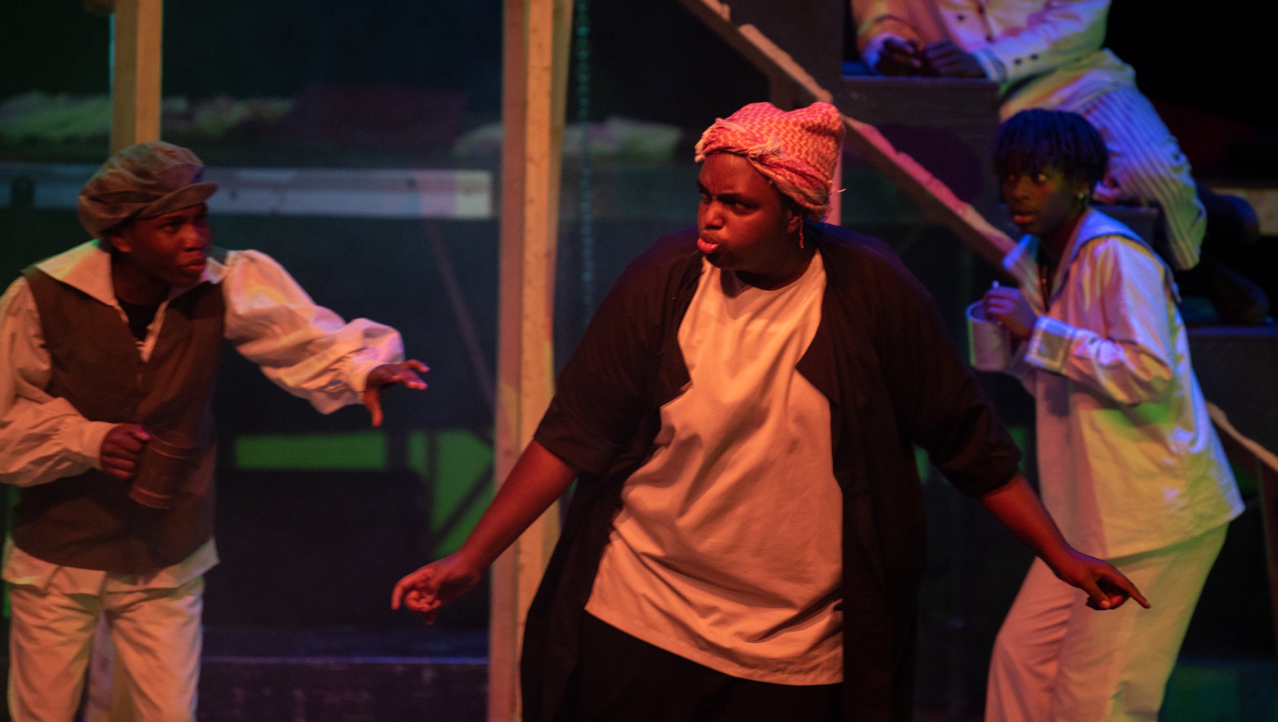 Three actors performing on stage in costume; one woman in the center with a headscarf, another woman on the left in a cap, and a woman on the right with dreadlocks, set against a dark background with theatrical lighting.