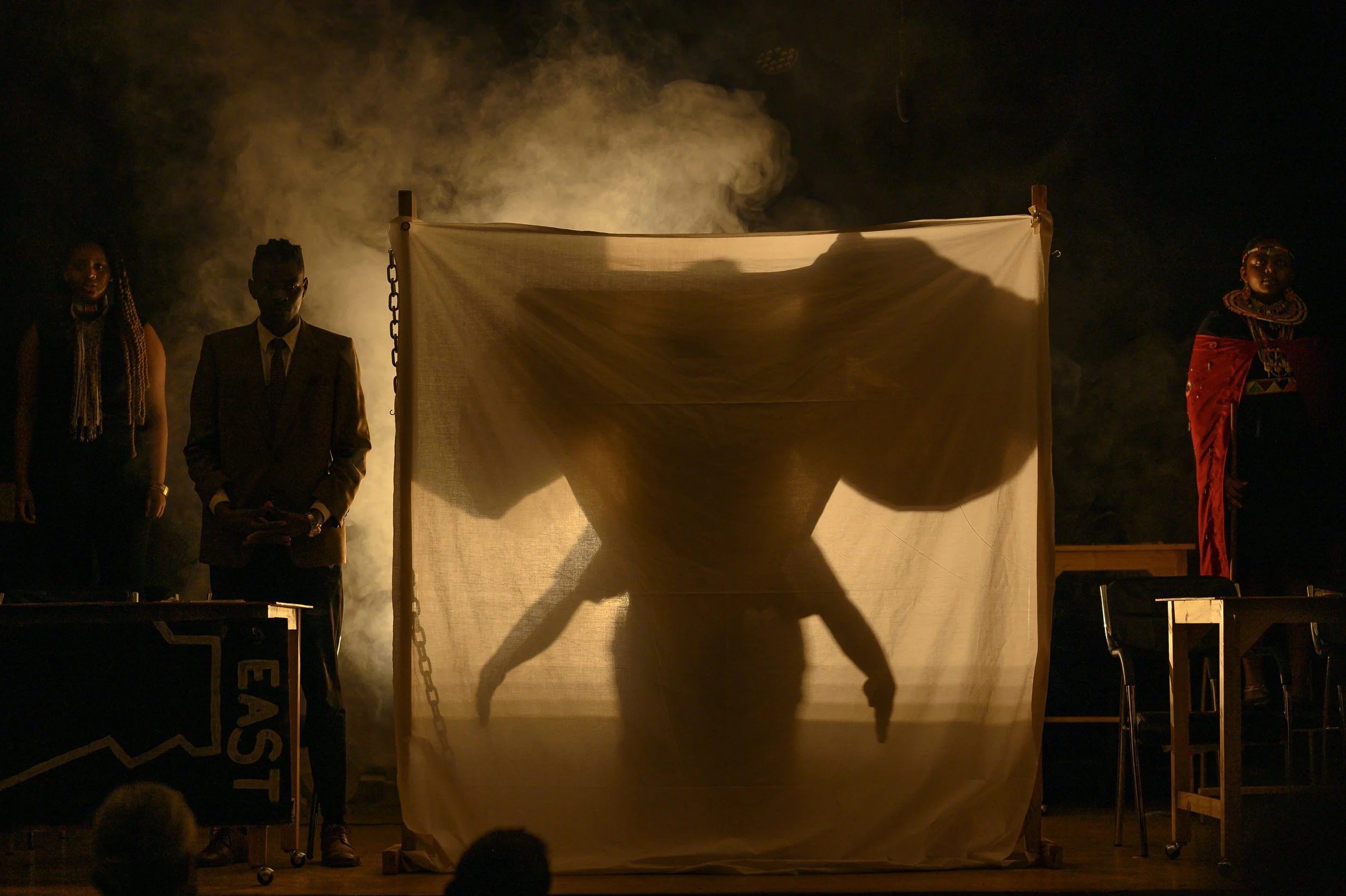 A theatrical performance with a silhouette of a person in a headstand pose behind a semi-transparent curtain, flanked by two individuals dressed in traditional attire, with smoke and dramatic lighting.