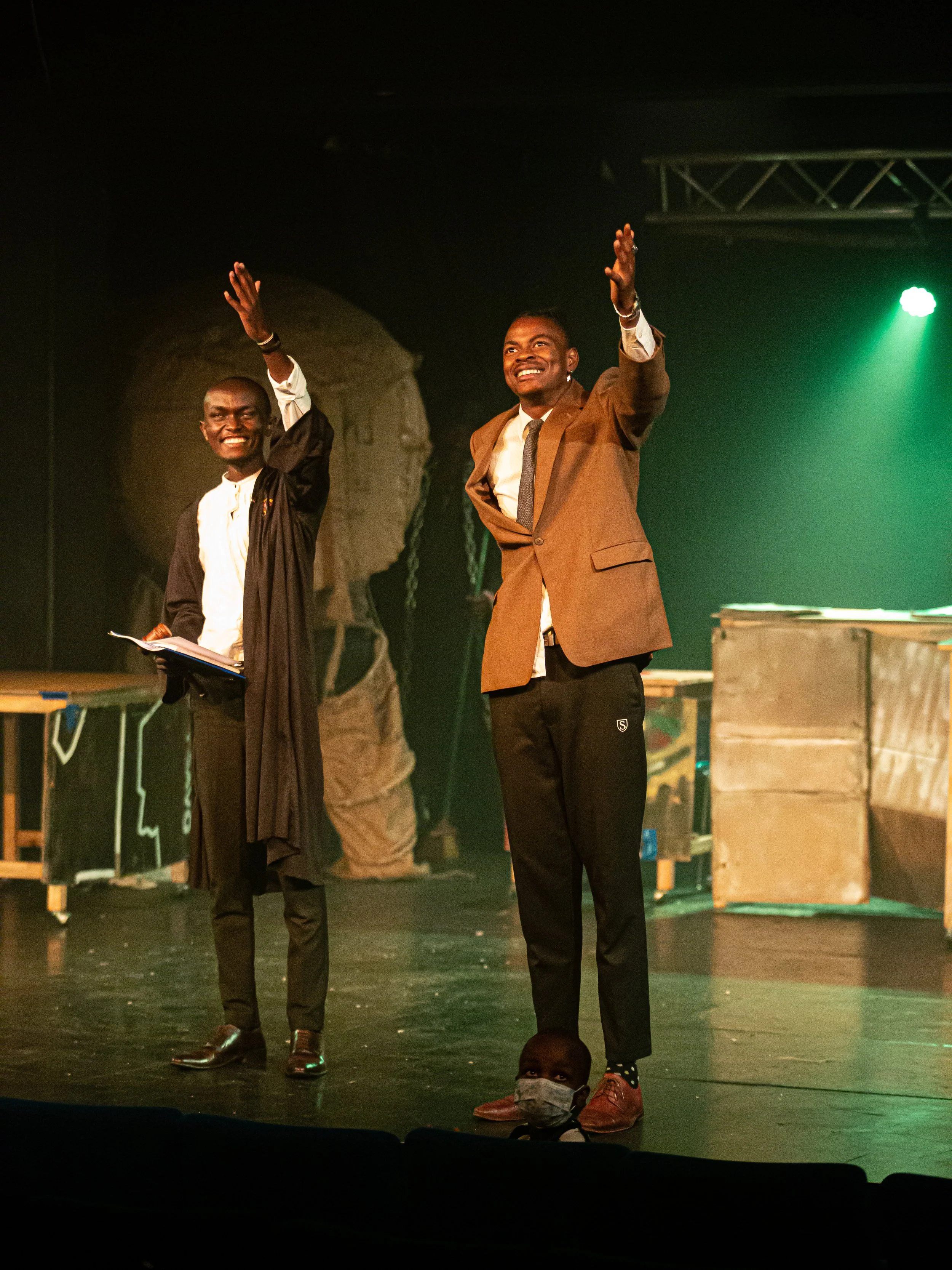 Two men on stage, one in a graduation gown and the other in a suit, waving to the audience, smiling, with stage lights and equipment in the background.